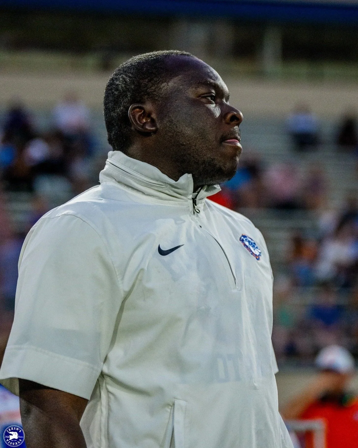 A man with dark skin and short dark hair, wearing a light gray sports jacket with the Nike logo and a patch that has a blue, white, and red design, standing outdoors during the daytime.
