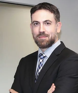 A man with a beard and short dark hair, wearing a black suit, light blue shirt, and checkered tie, standing with arms crossed against a neutral background.