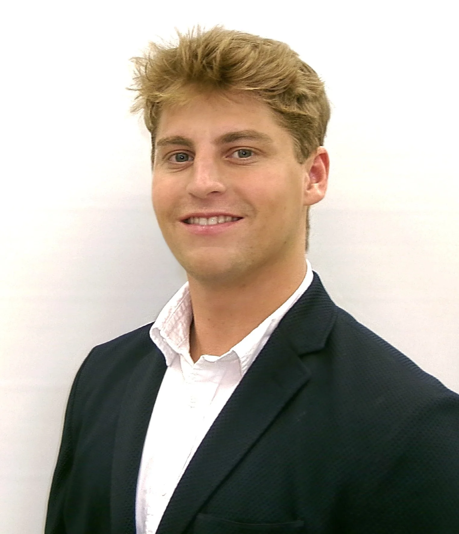 A young man with blonde hair, wearing a white collared shirt and blue suit jacket, smiling against a plain white background.