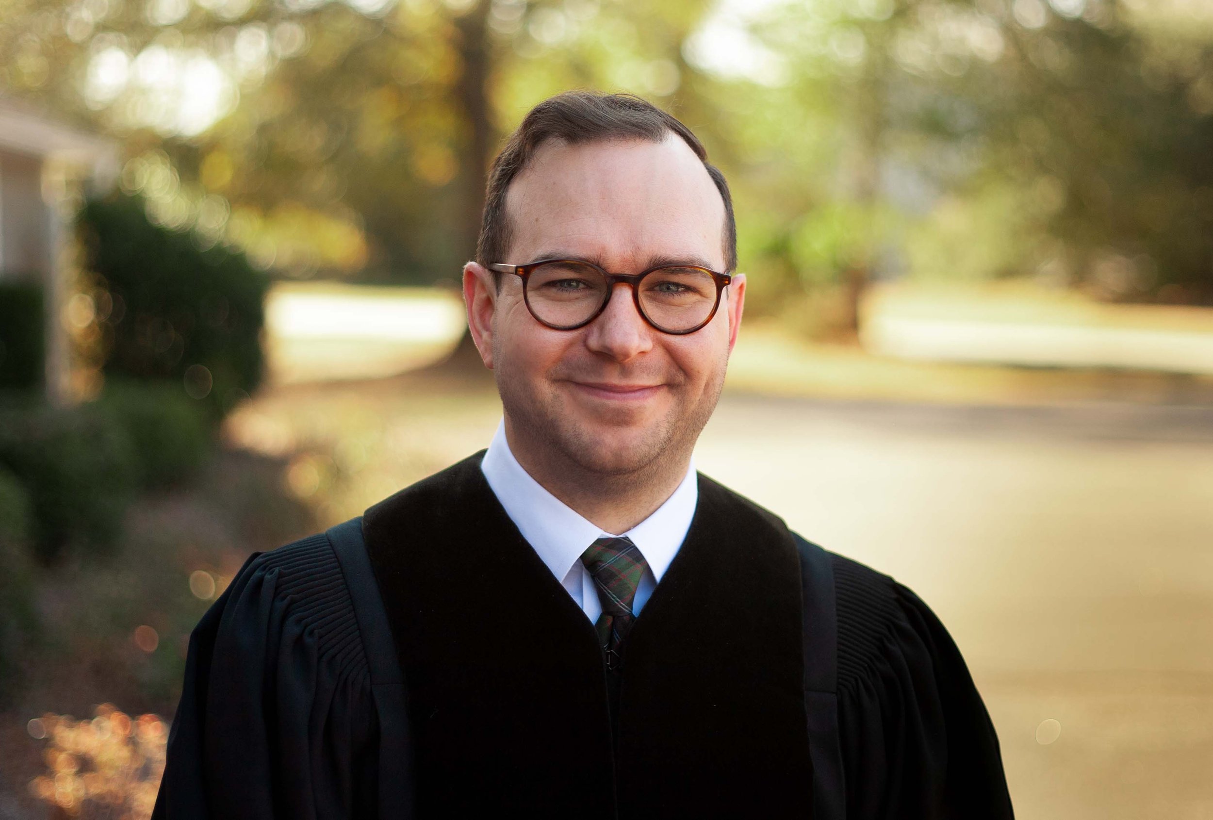 A man in graduation attire, wearing glasses and a tie, smiling outdoors with blurred trees and autumn foliage in the background.