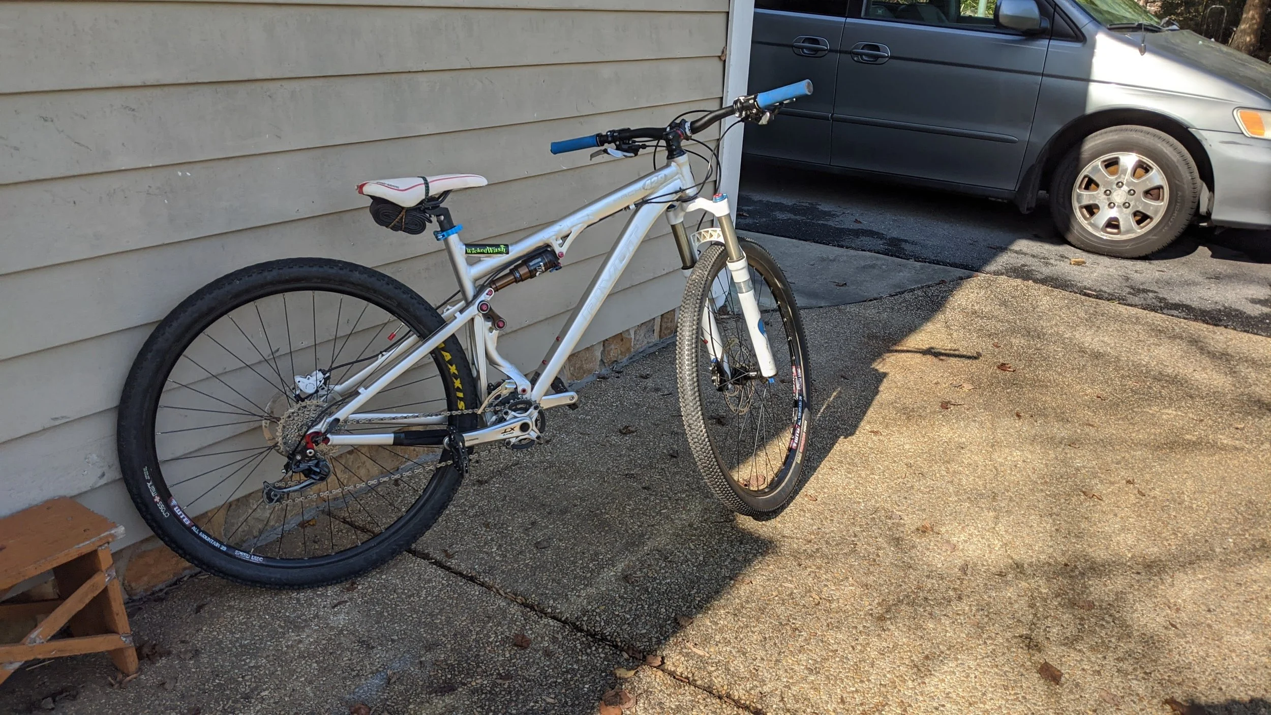 White mountain bike leaning against the beige house siding, with a gray car parked on the street in the background, on a sunlit sidewalk.