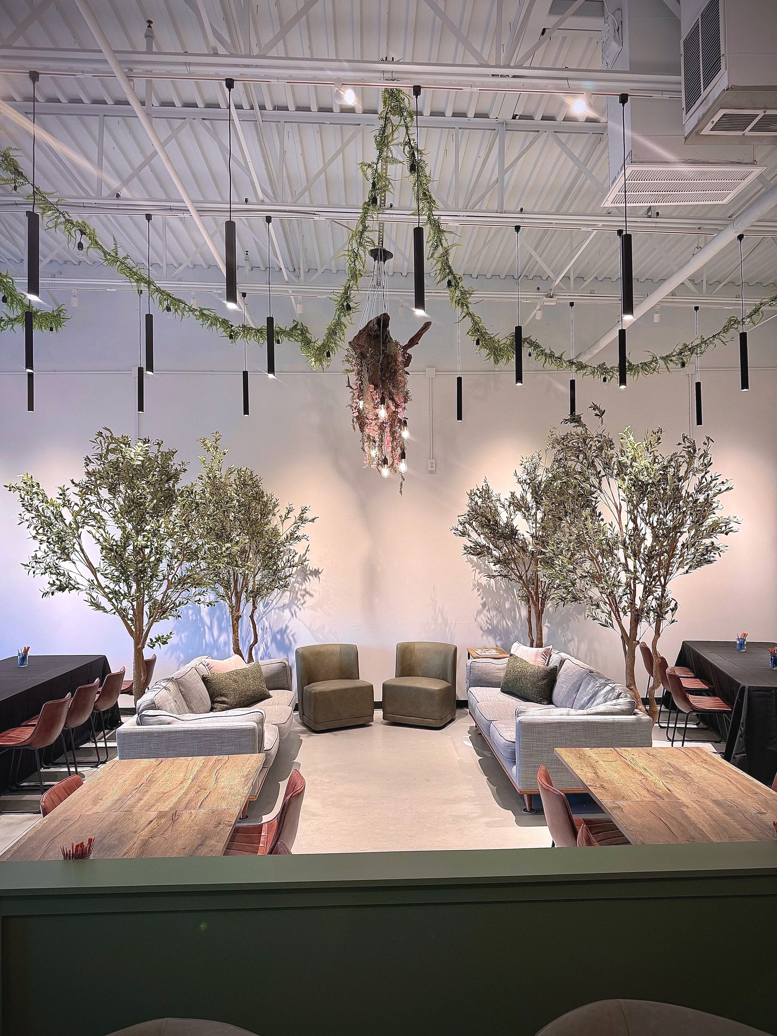 Soft seating lounge area at The Painted Plant art studio in Spring Hill, TN, featuring grey sofas, olive green chairs, tables, and decorative greenery for relaxing or playing a family game while projects finish.