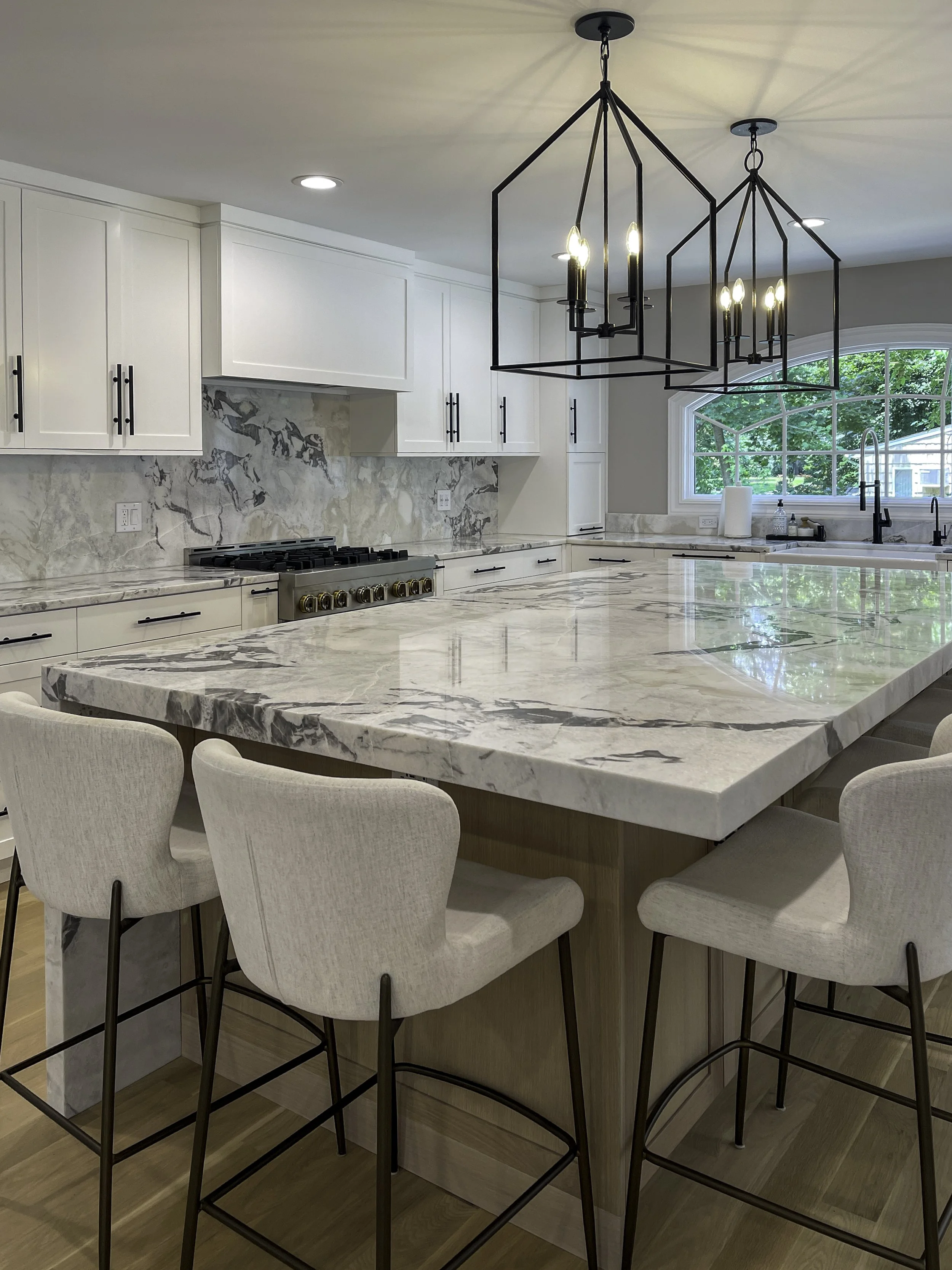 Kitchen island with waterfall marble countertop, upholstered stools, and black lantern-style pendant lighting in Plandome, NY