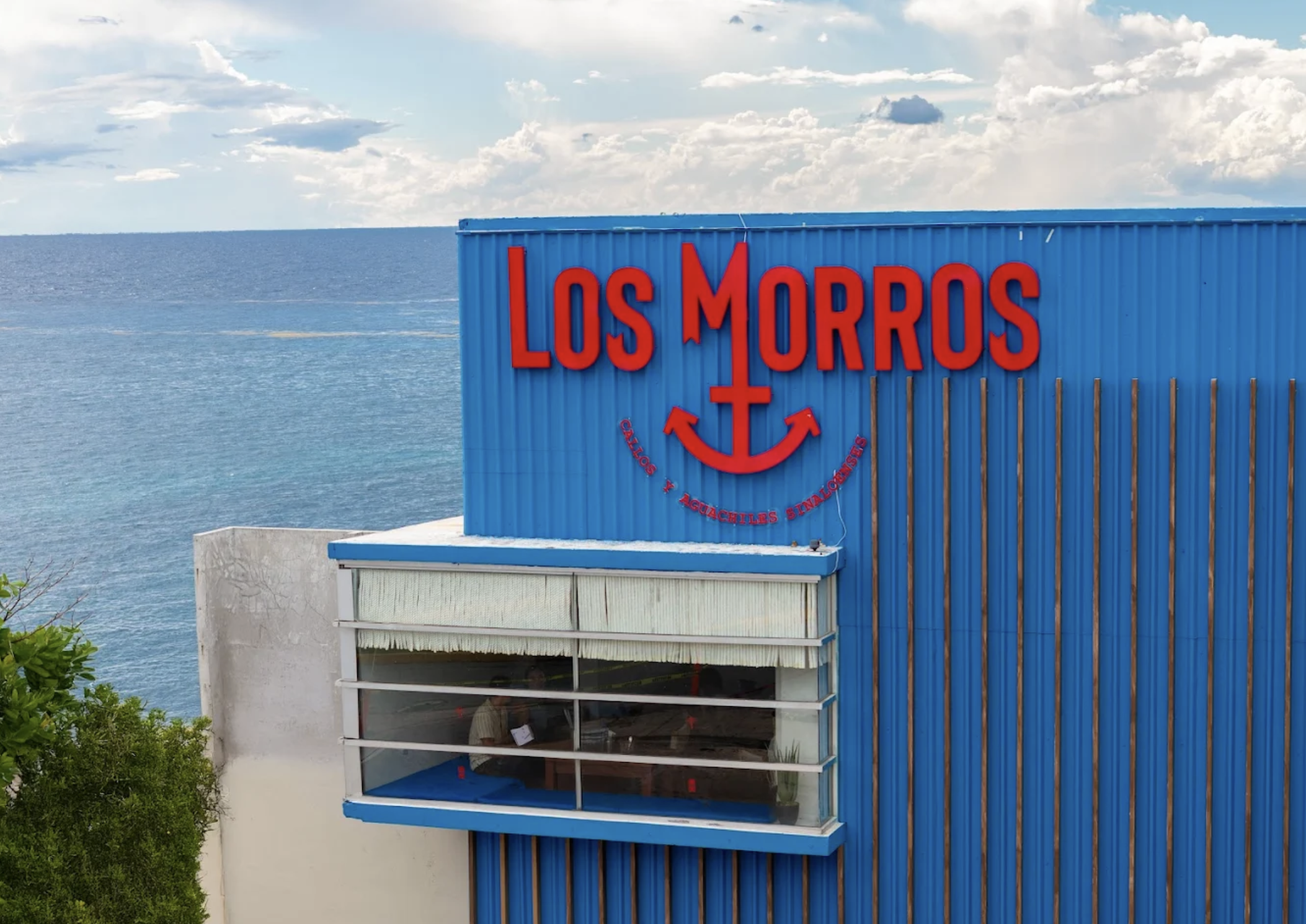 Sign with the words 'Los Morros' and an anchor symbol underneath, with the phrase 'Calzos y Aguachiles Sinaloenses' below. The building is blue, located near the water, with an ocean in the background under a partly cloudy sky.
