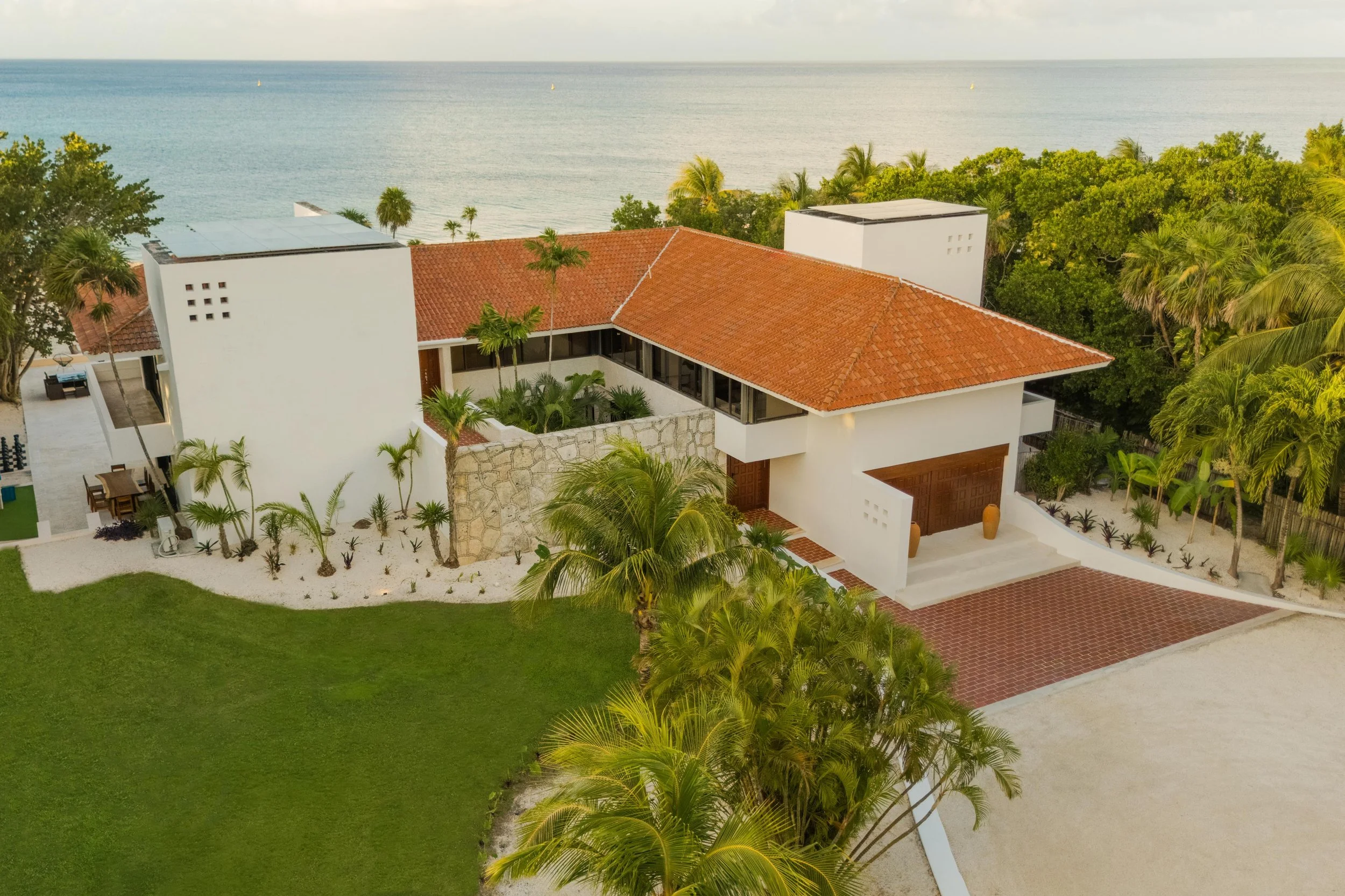 Aerial view of a modern house with a red tile roof, surrounded by lush green tropical trees and a well-maintained lawn, overlooking the ocean in the background.