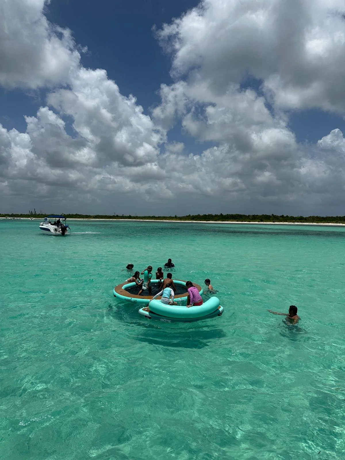 People enjoying a day on a floating water trampoline in clear turquoise water near the coastline, with a boat in the distance and a cloudy sky overhead.