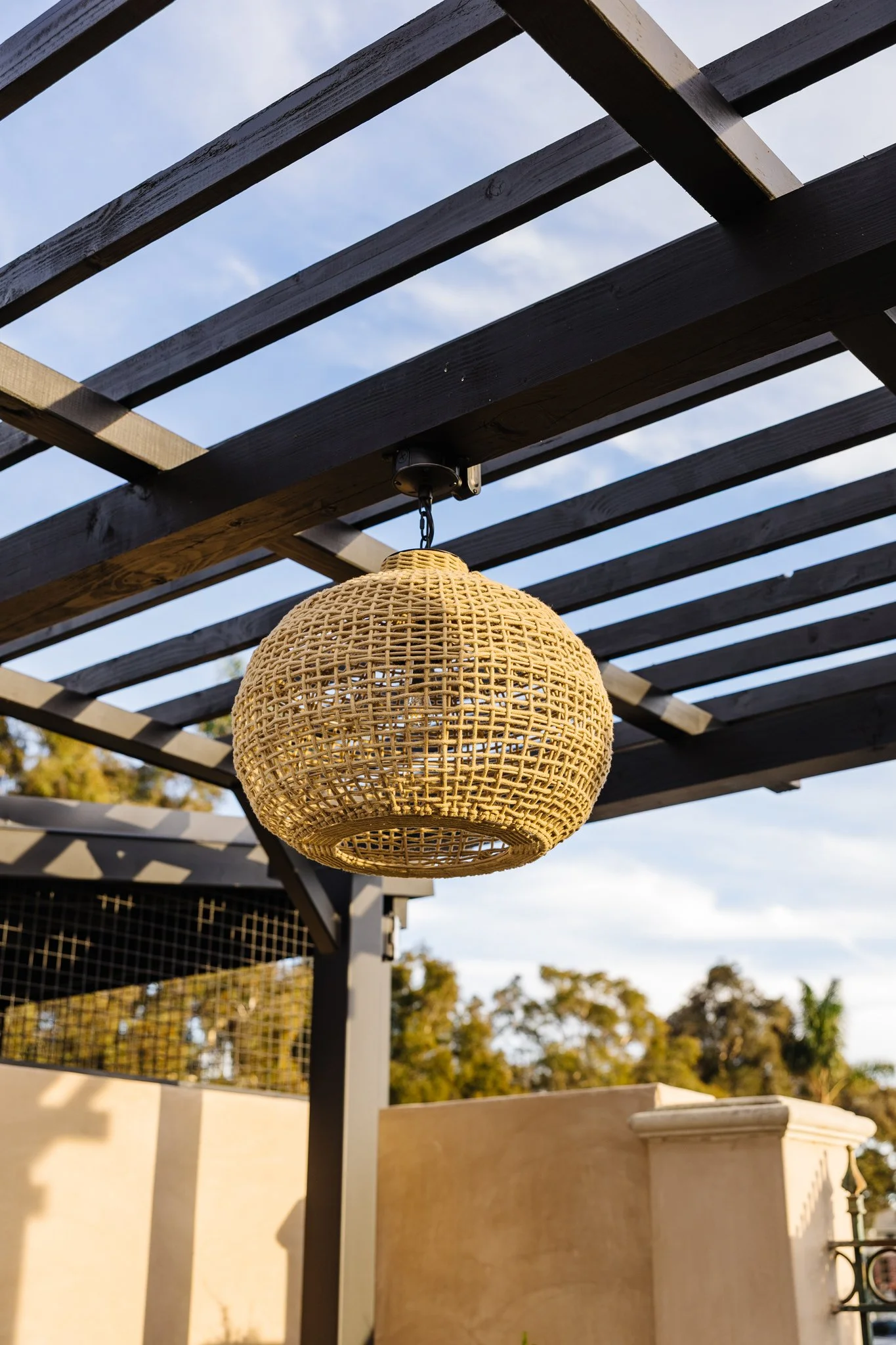 A woven pendant light adds a soft glow beneath the pergola, creating warmth and atmosphere as day turns to night. Its natural texture balances the outdoor setting while providing intentional lighting for gatherings below.