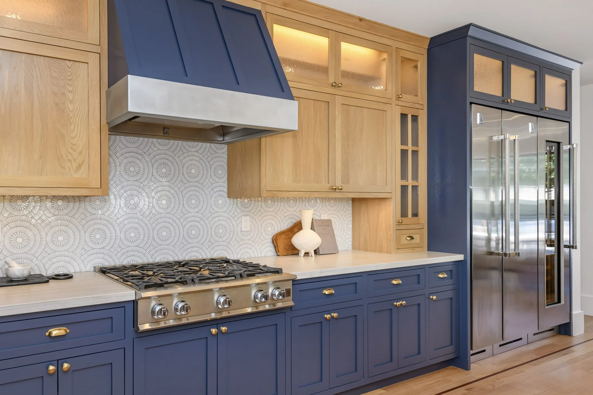 Kitchen with blue and natural wood cabinets, gray patterned backsplash, stainless steel range hood, gas stove, and large stainless steel refrigerator.