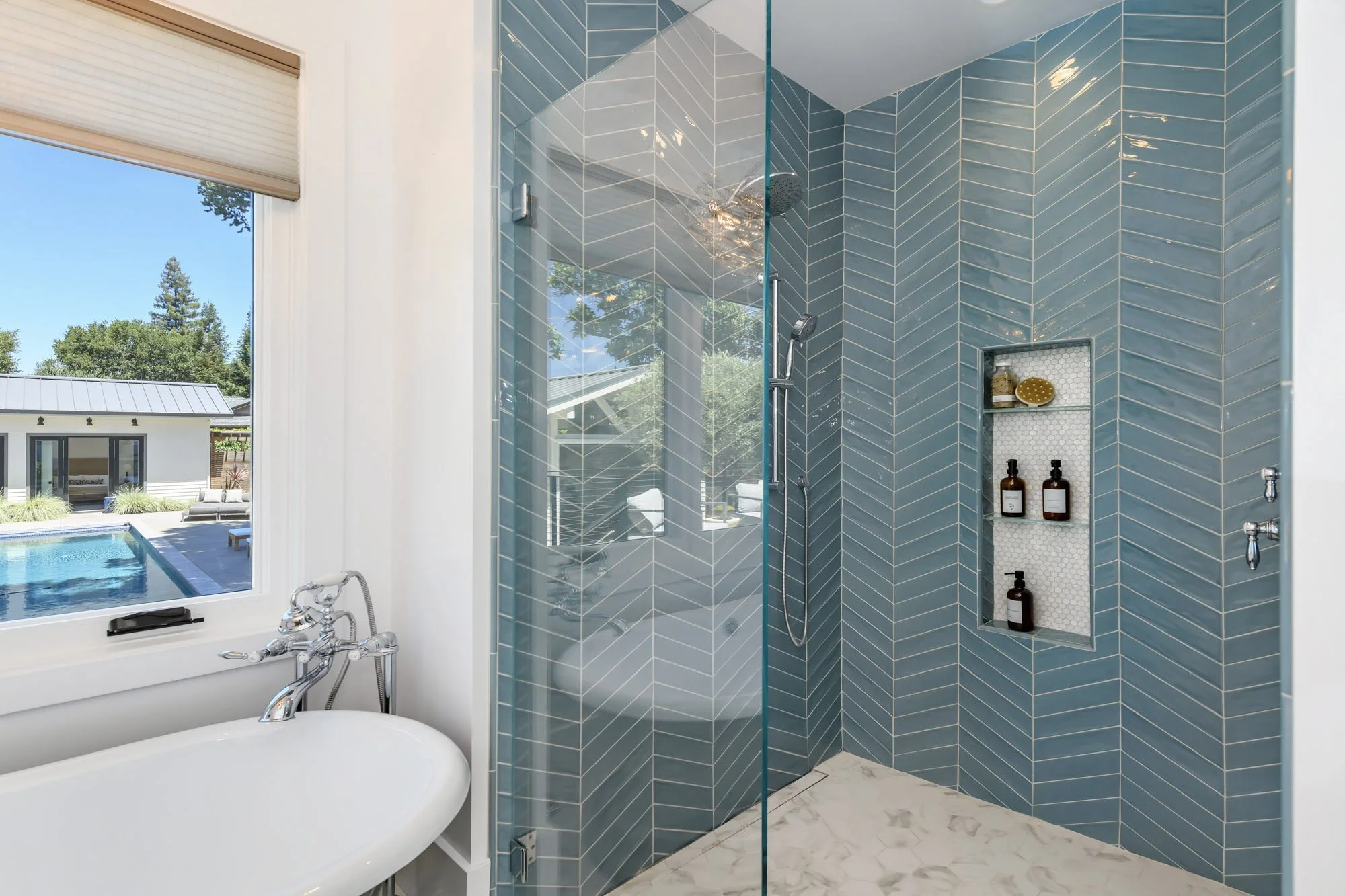 Bathroom with a bathtub beneath a window, and a glass-enclosed shower with blue subway tile walls, built-in niche, and toiletries.
