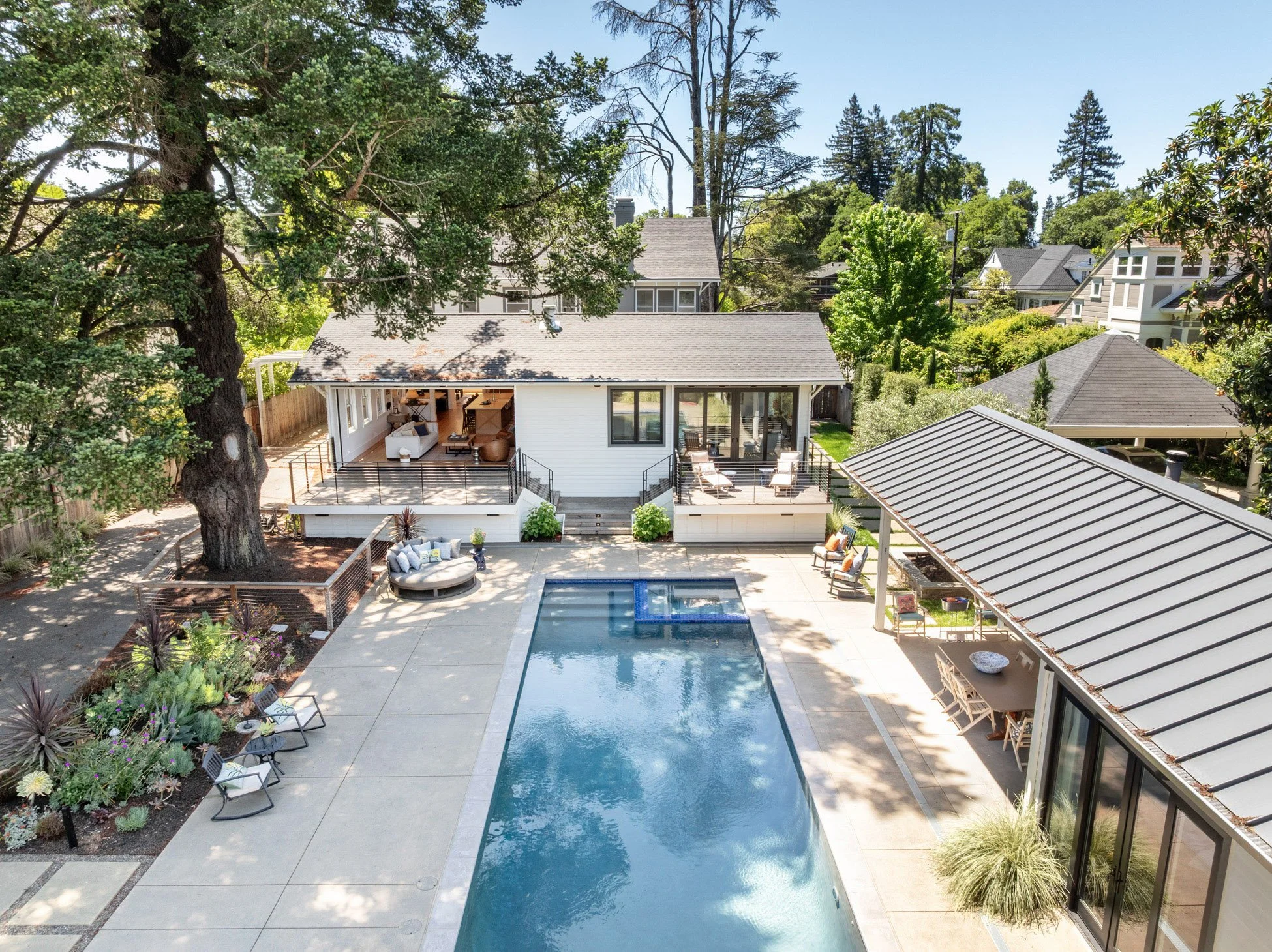 A backyard with a swimming pool, outdoor seating, and a white house with a deck, surrounded by trees and other houses.