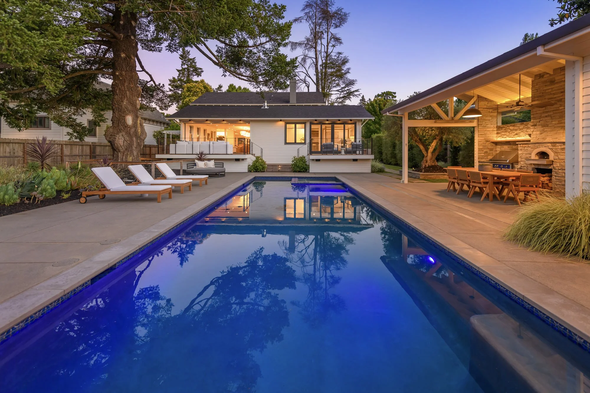 A backyard with a swimming pool, lounge chairs, a covered patio with a dining area, and a modern house with large windows, all illuminated during twilight.