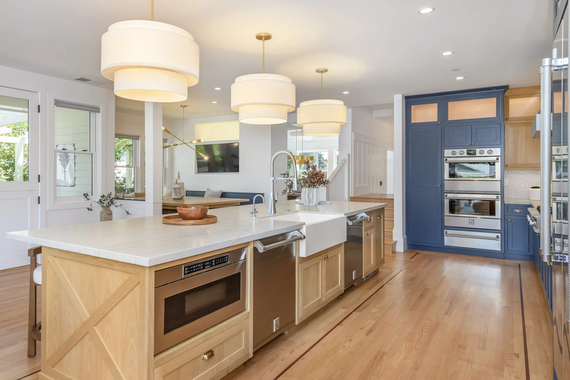 Modern kitchen with light wood cabinets, a large island with a white countertop, blue cabinets around the oven, and three large pendant lights hanging from the ceiling.