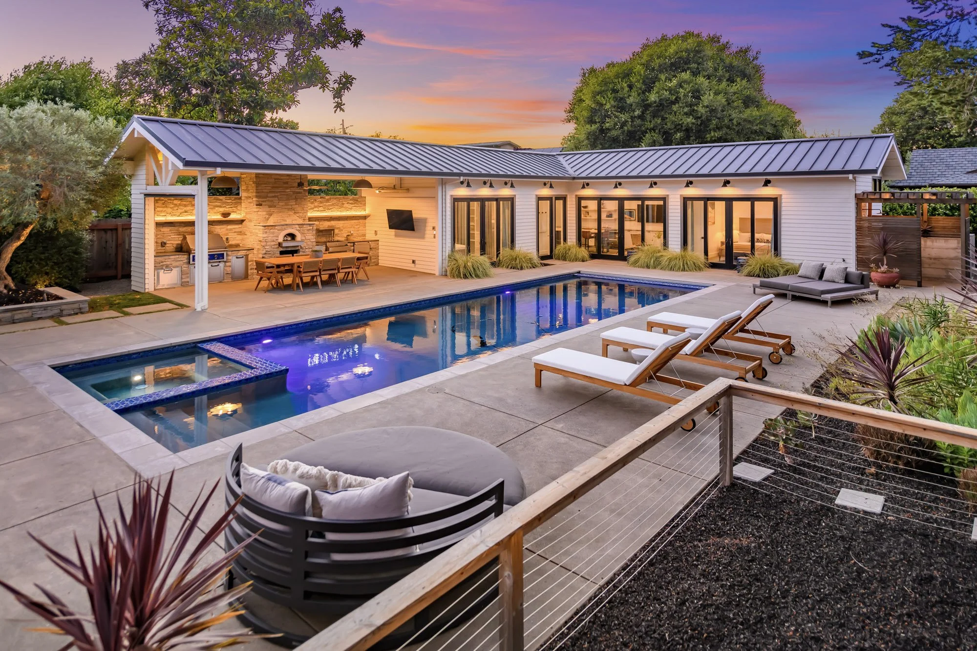 A backyard with a modern pool, outdoor seating, and a covered outdoor kitchen area at sunset.