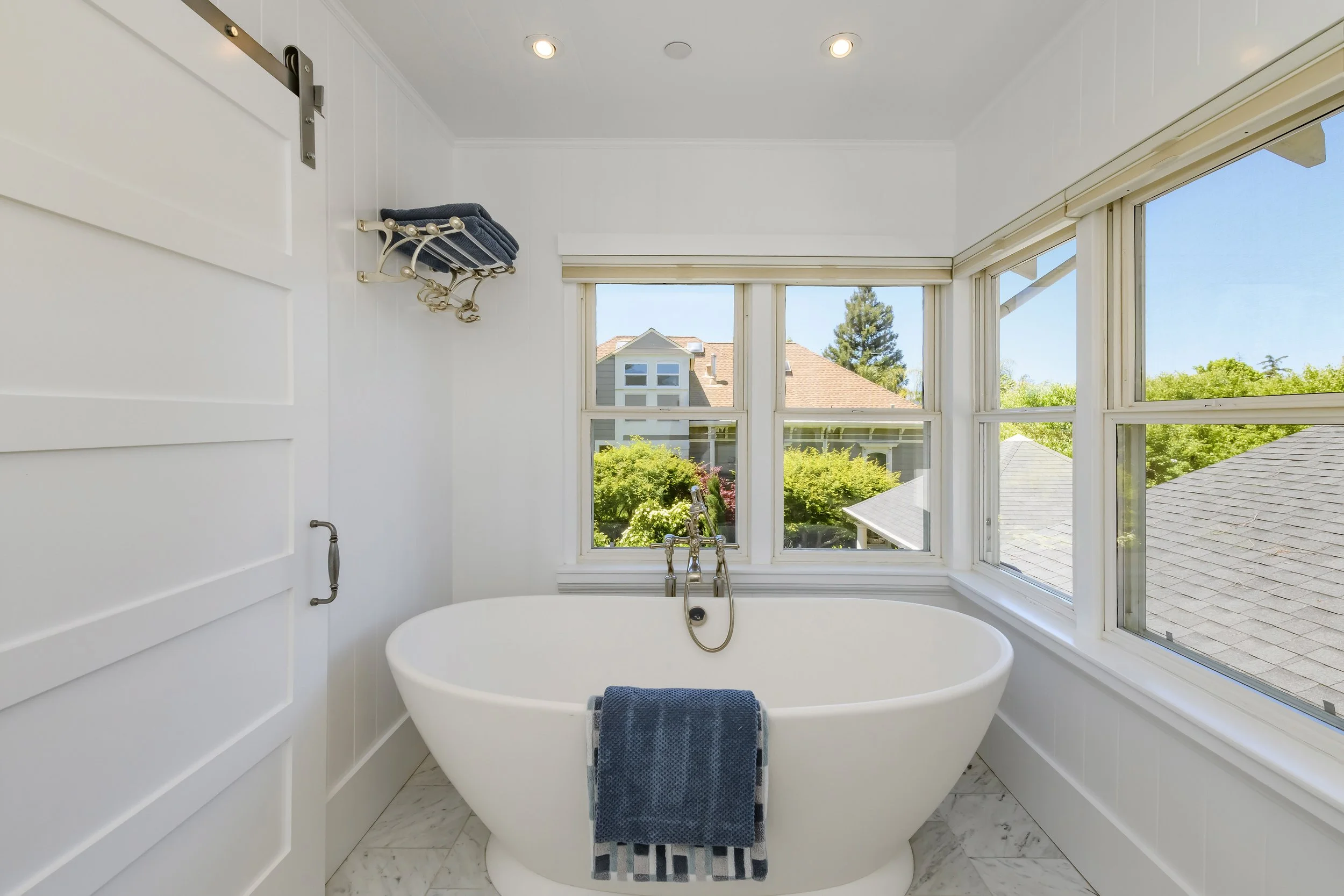 Bright bathroom with a white bathtub positioned in front of large windows showing a view of a house with a tile roof, trees, and a blue sky.