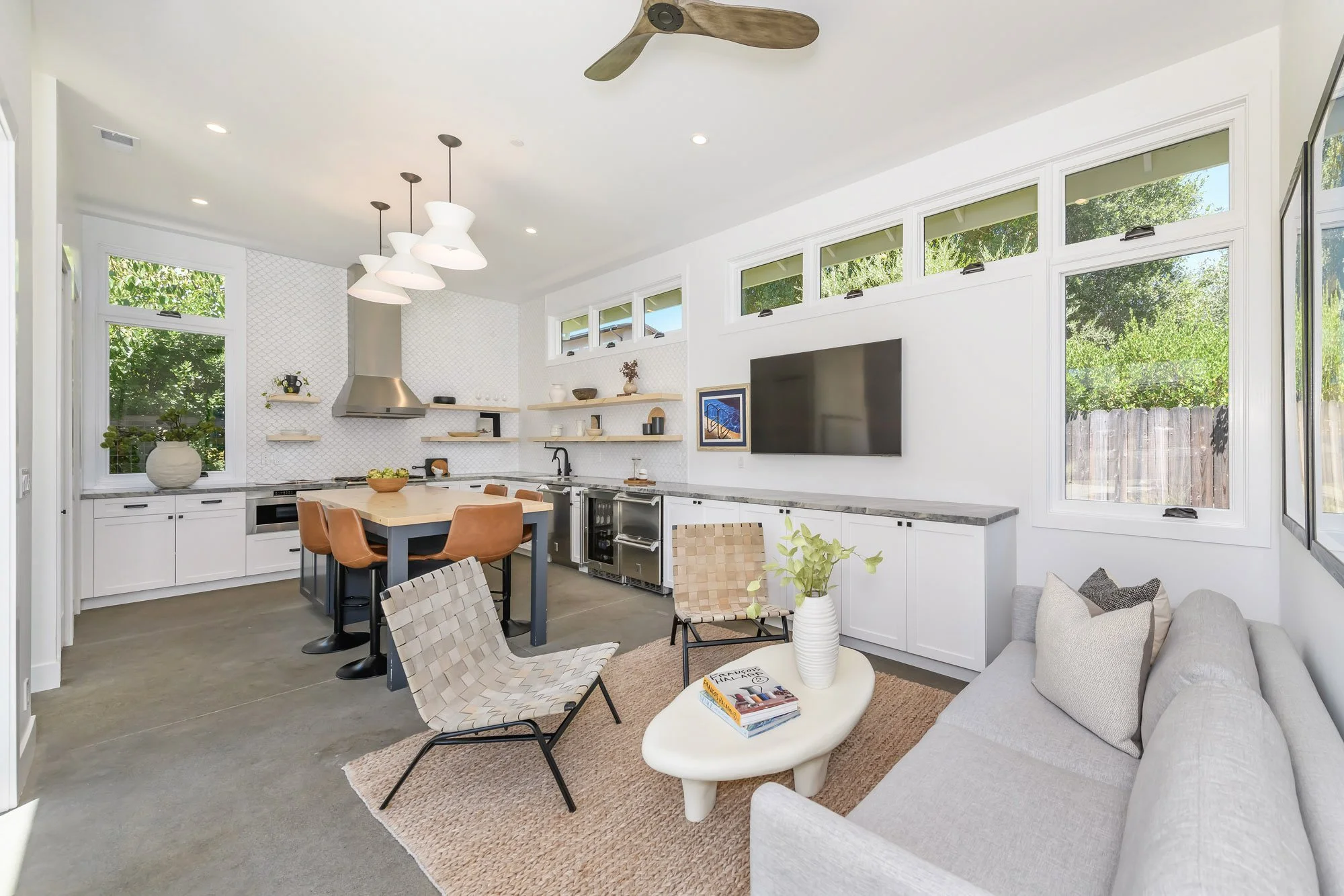 Modern open-plan living room and kitchen with white cabinetry, wooden shelves, a tiled backsplash, a wall-mounted television, and brown chairs, with large windows letting in natural light.