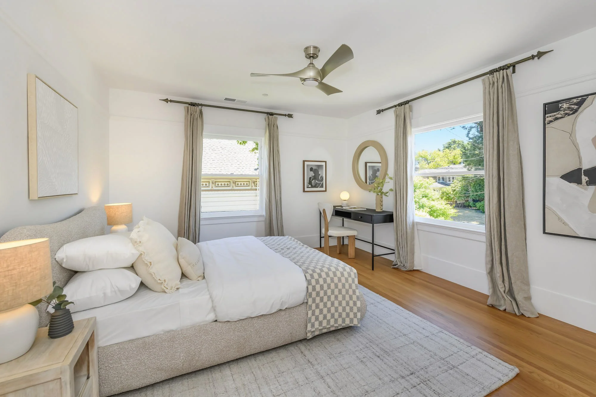 Bright bedroom with a bed, nightstand, two table lamps, a small desk with a chair, mirror, and large windows with beige curtains, hardwood flooring, and artwork on the walls.