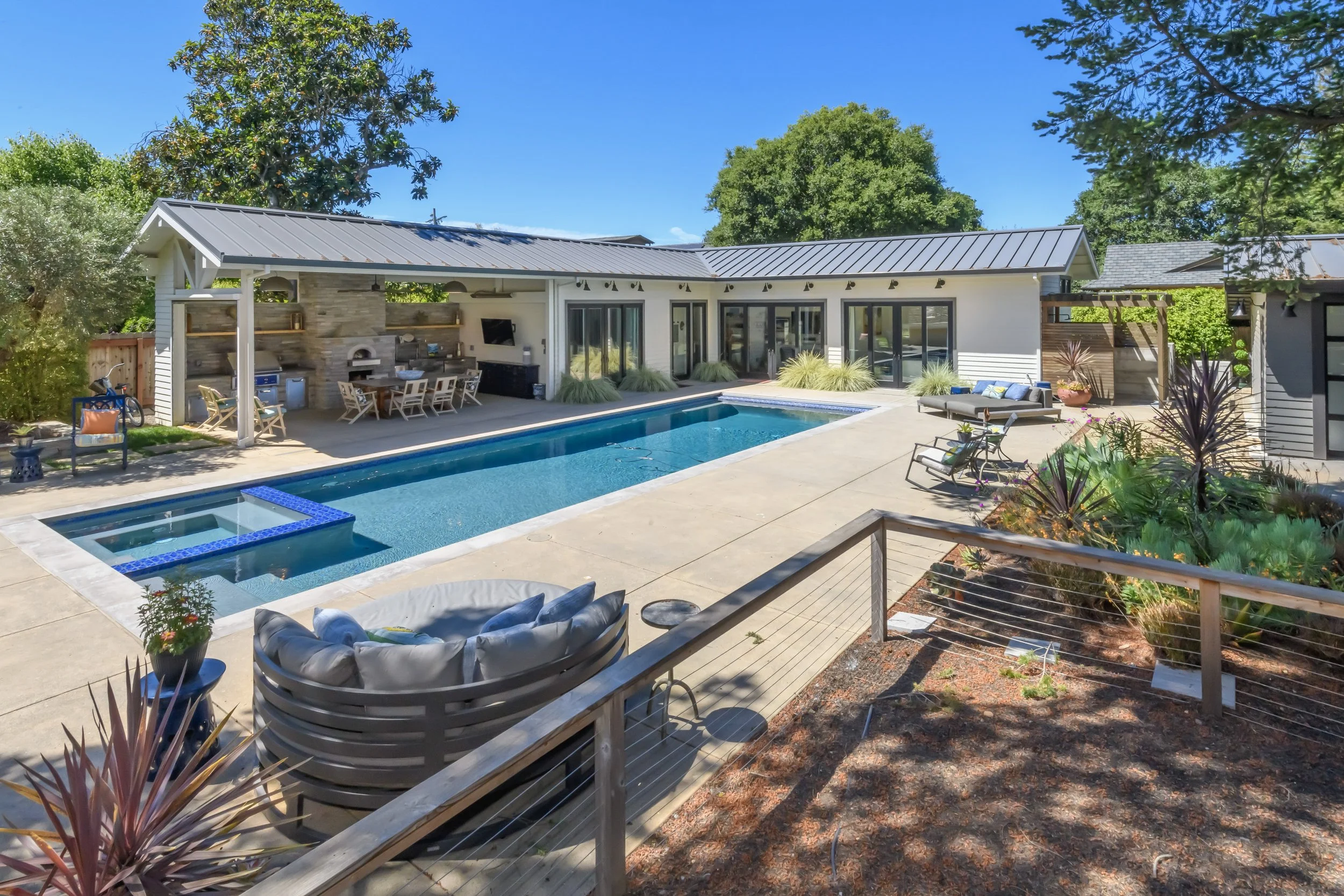 Modern backyard with a rectangular pool, hot tub, outdoor seating, and a covered kitchen area, surrounded by trees and landscaping.