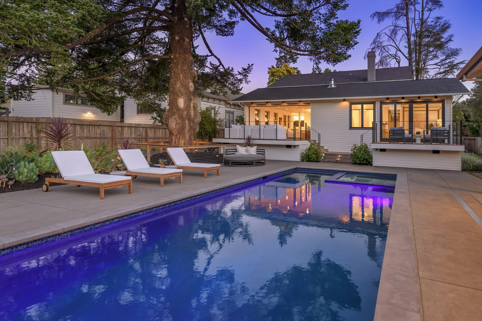 Backyard with swimming pool and outdoor seating, fence, large trees, illuminated house, evening sky
