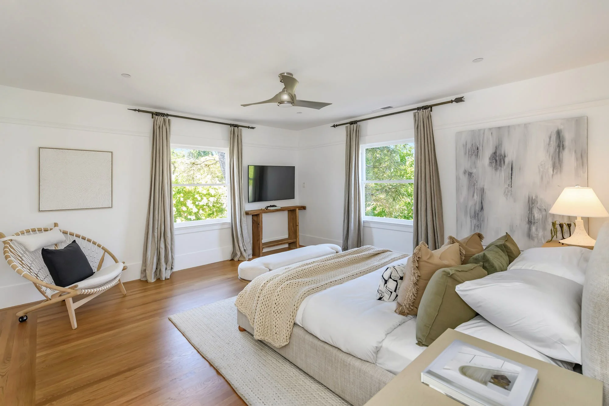 A cozy bedroom with a large bed, multiple throw pillows, a nightstand with a lamp, and a large abstract painting on the wall. There is a window with beige curtains and outside greenery visible. A chair with a pillow, a mounted TV, and light wooden fl