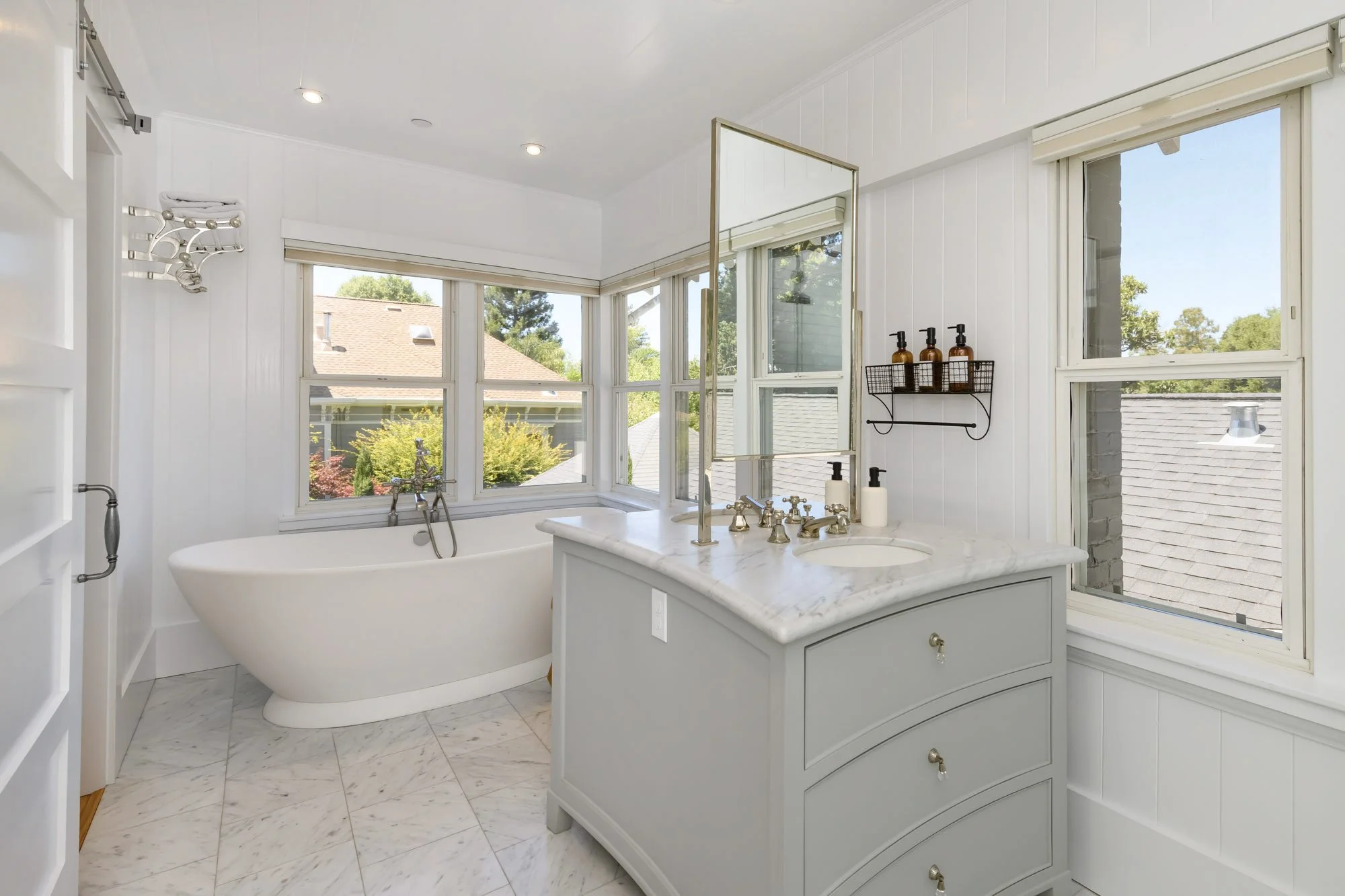 A bright bathroom with large windows, a white bathtub, a light gray vanity with a marble top, and a bathroom mirror.