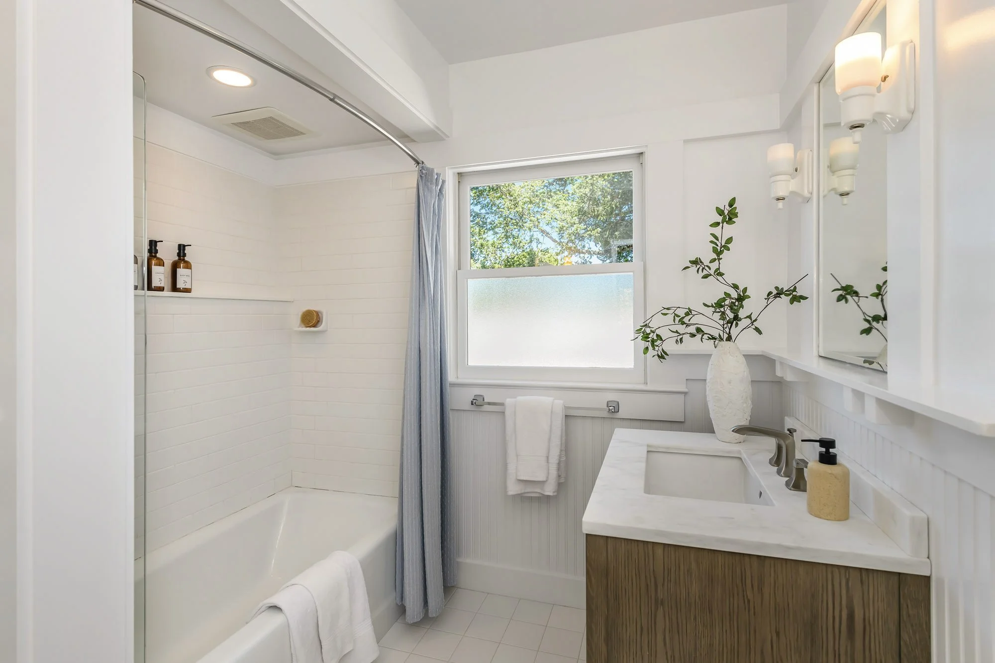A bright bathroom with a bathtub and shower, white tiled walls, a window showing green trees outside, a white towel on a horizontal bar, a marble vanity with a sink, a mirror, a vase with branches, and a soap dispenser.