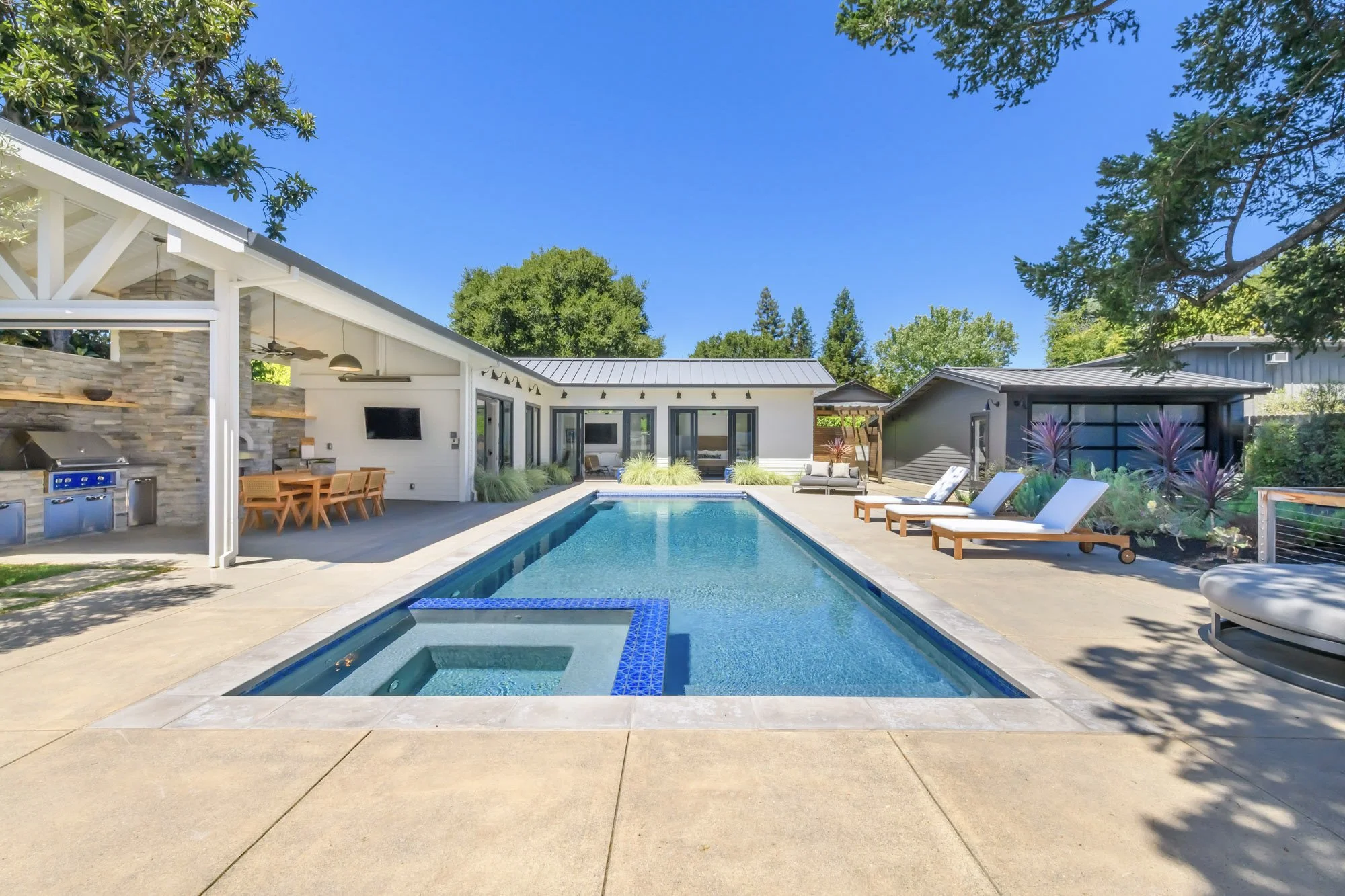 Modern backyard with rectangular swimming pool, multiple lounge chairs, outdoor dining area, and a covered outdoor kitchen under a bright blue sky.