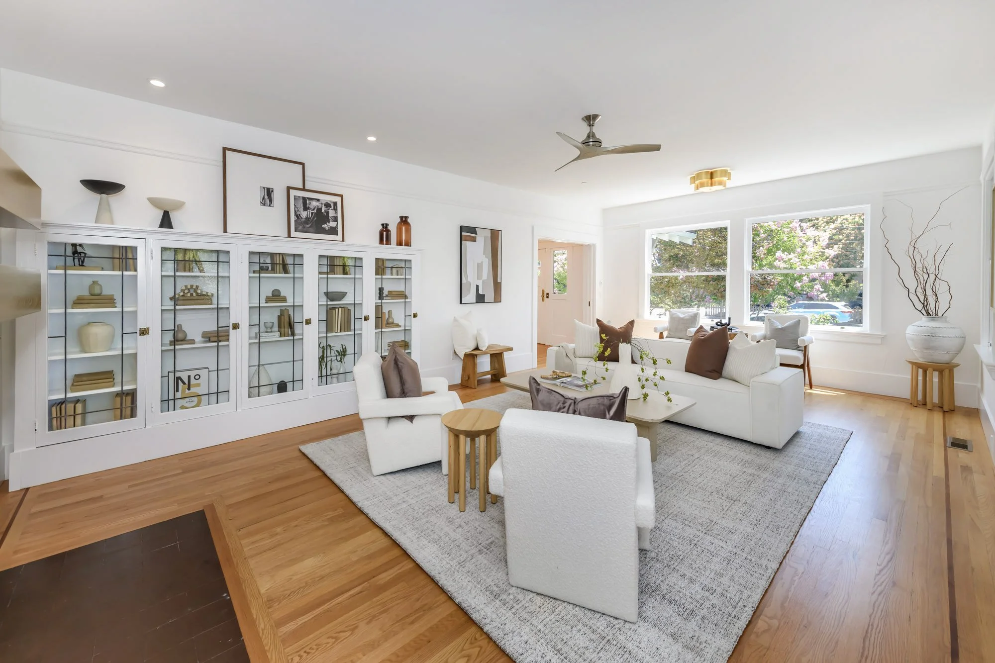 Bright living room with white walls, wooden floors, and large windows with views of greenery. Furnished with a white sofa, armchairs, and pillows, centered around a low coffee table on a gray rug. Contains built-in glass-fronted display cabinets, art