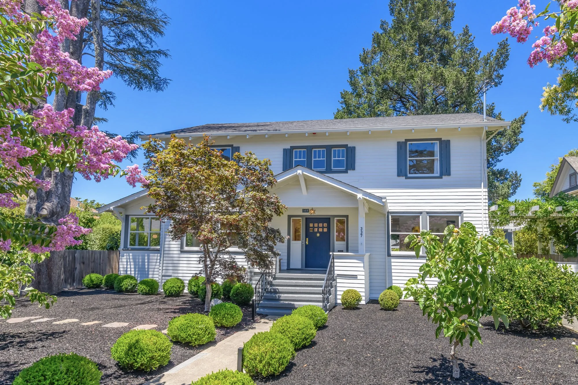 White two-story house with blue front door, surrounded by green bushes and trees, under a clear blue sky.