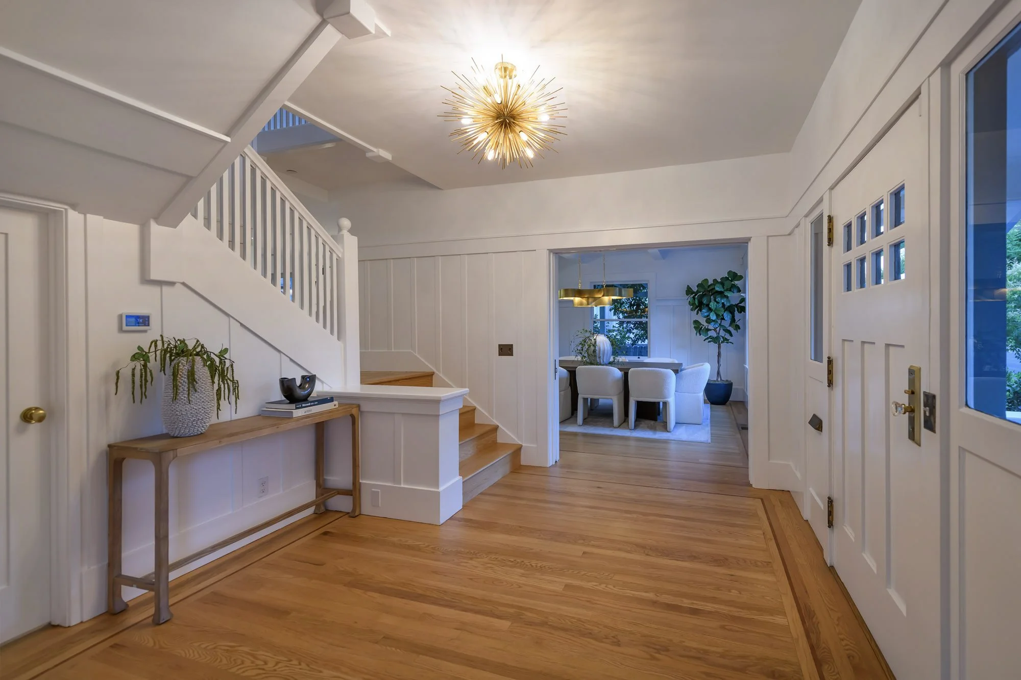 Entryway with wooden flooring, white walls, staircase on the left, a console table with a plant, and a view into a dining room with white furniture and a large window.