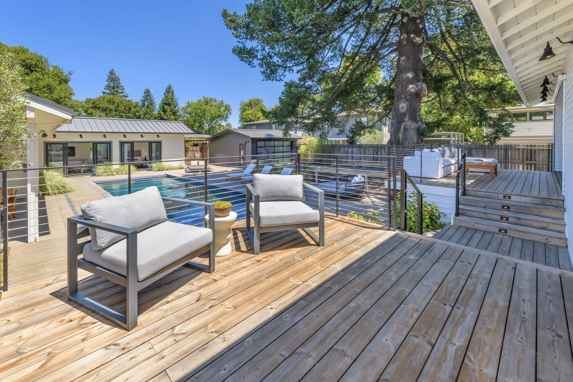 Outdoor wooden deck with seating area, overlooking a swimming pool surrounded by modern houses, large trees, and clear blue sky.