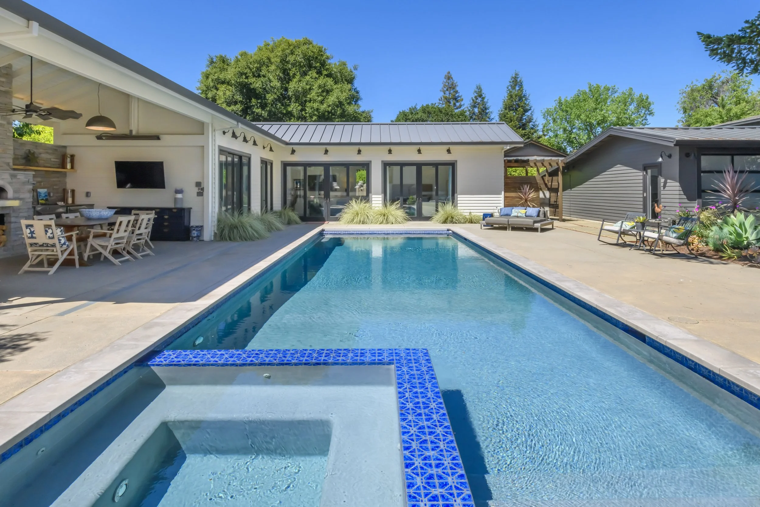 Backyard with a modern swimming pool, patio seating, and outdoor dining area under a clear blue sky.