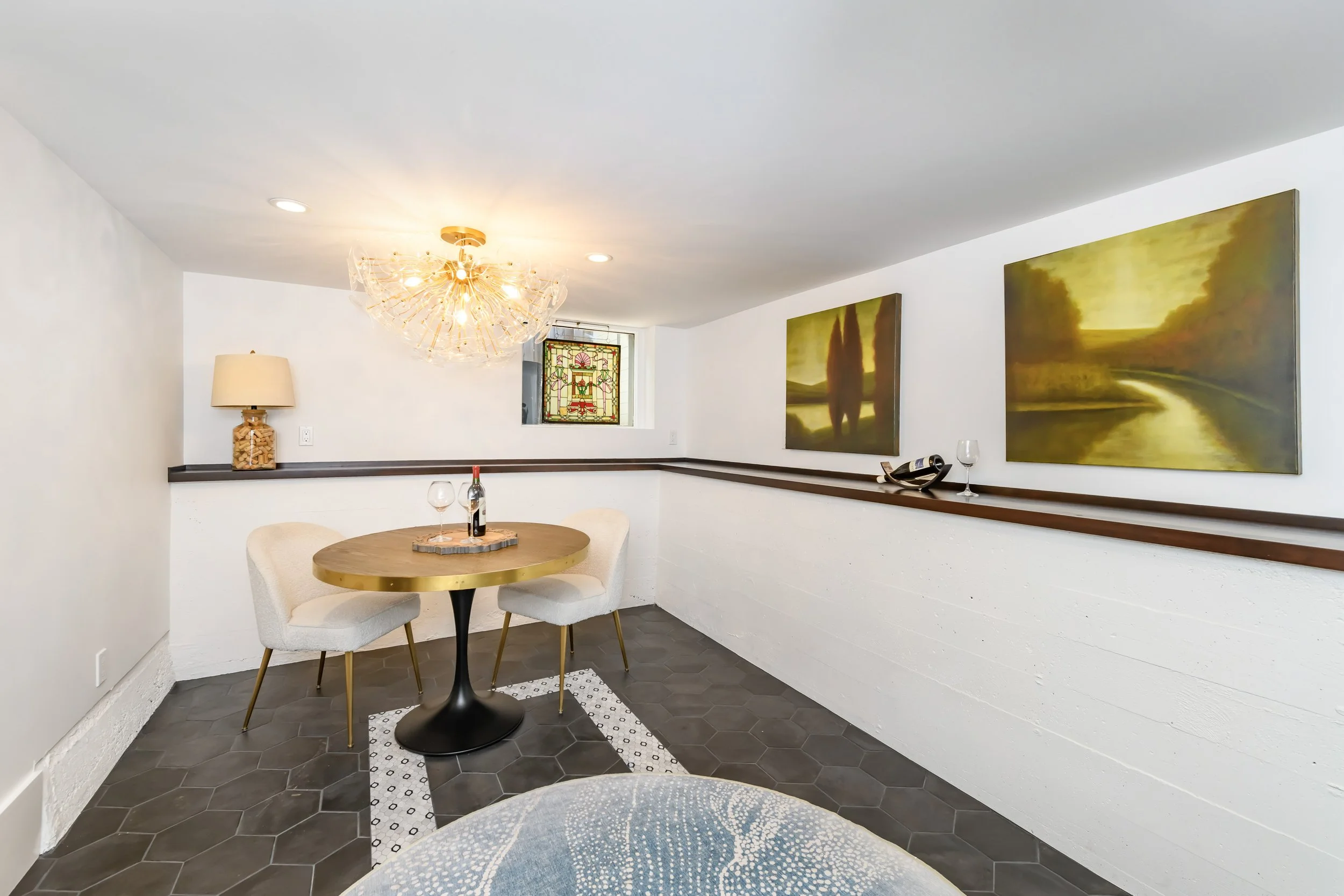 A small dining nook with a round table, two plush chairs, wall art, a small stained glass window, and a modern chandelier in a cozy room.