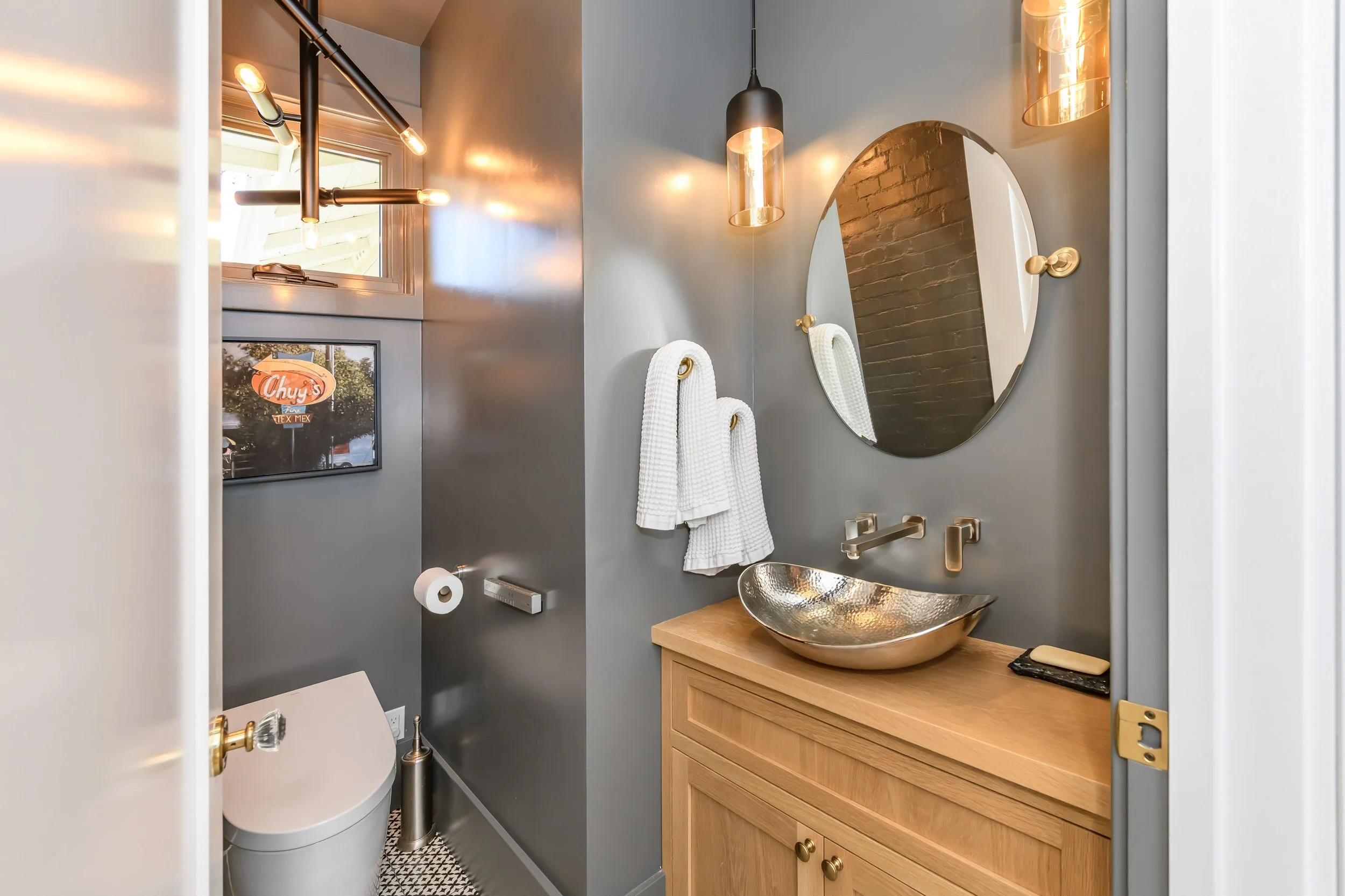 Small modern bathroom with a wood vanity, metallic vessel sink, oval mirror, black pendant lights, gray walls, and a wall-mounted toilet.