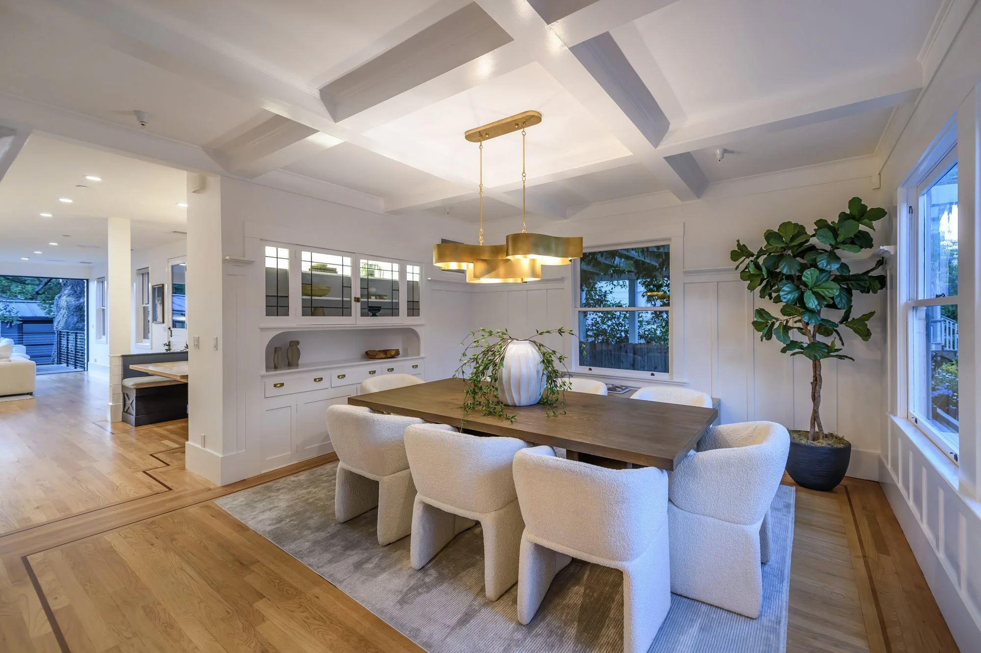 Dining room with a wooden table, eight white upholstered chairs, a large gold pendant light, a potted fiddle leaf fig tree, and windows showing outdoor greenery.