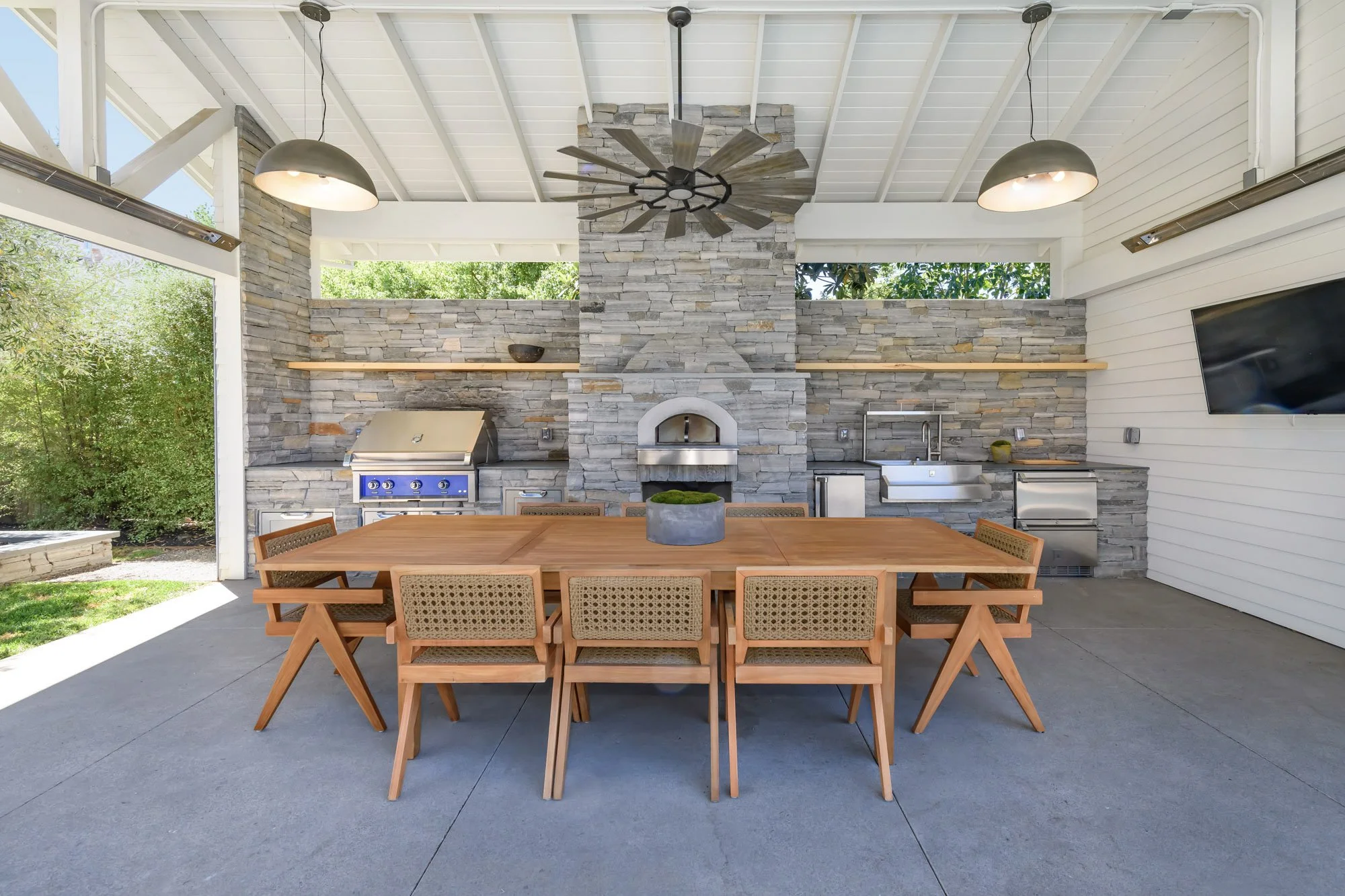 Outdoor patio with a large wooden dining table, eight chairs, stone fireplace, built-in grill, sink, TV, ceiling fans, and sliding doors.