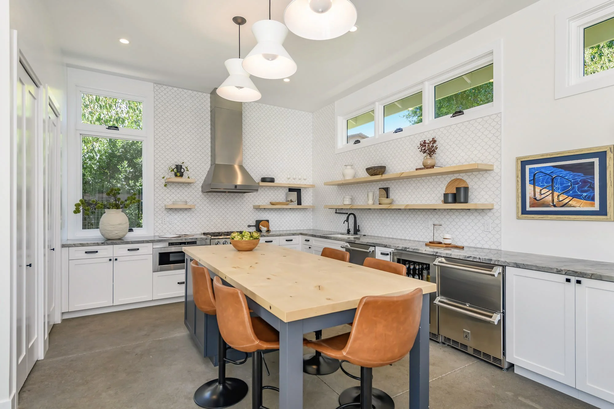 Modern kitchen with white cabinets, a wooden island, brown chairs, open shelves, stainless steel appliances, a window with greenery outside, and a colorful artwork on the wall.