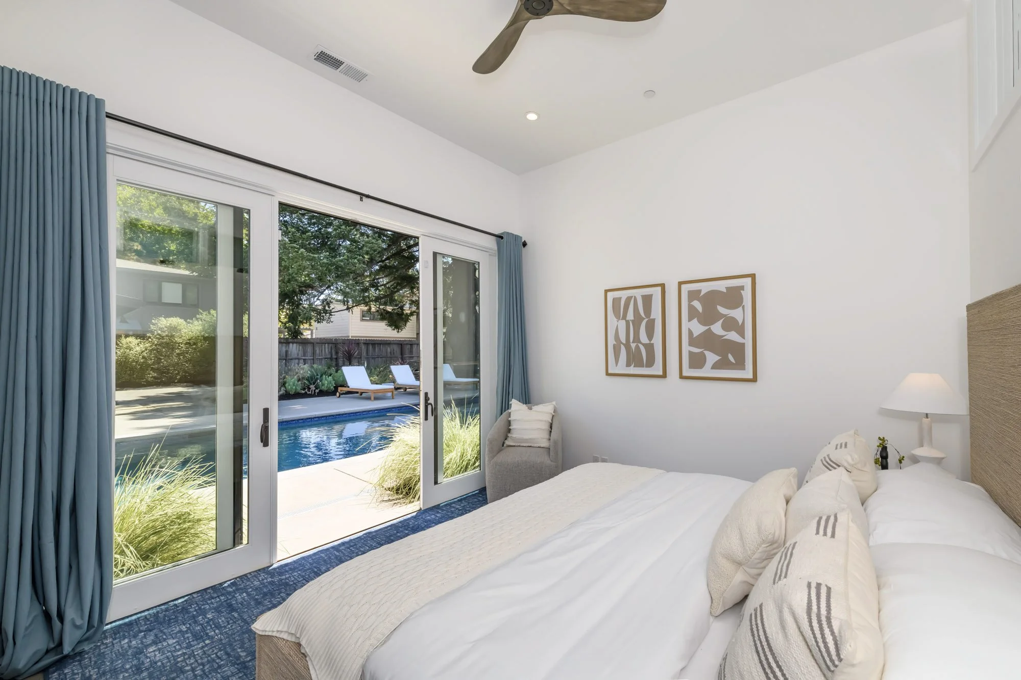 A bedroom with a large sliding glass door leading to a backyard with a pool and lounge chairs. The room has a bed with white bedding, a side table with a lamp, and two framed abstract art pieces on the wall. Blue curtains frame the glass door.
