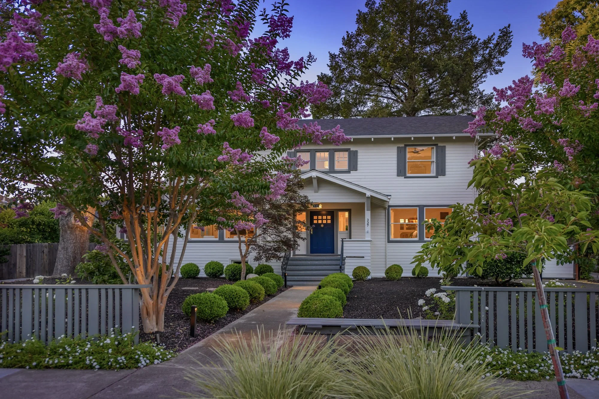 A two-story house with white siding, blue front door, and gray window shutters, surrounded by flowering trees and a landscaped front yard at dusk.
