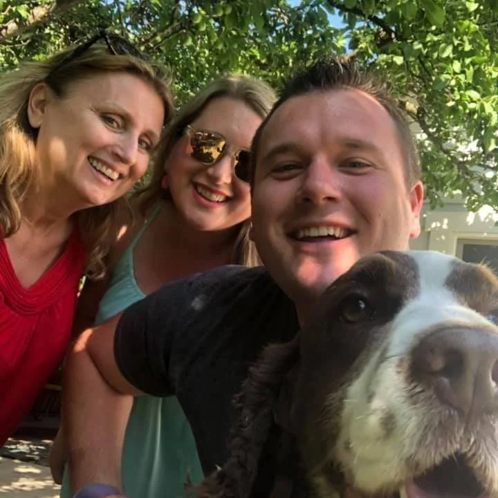 A group of three smiling people and a dog outdoors under green tree branches.