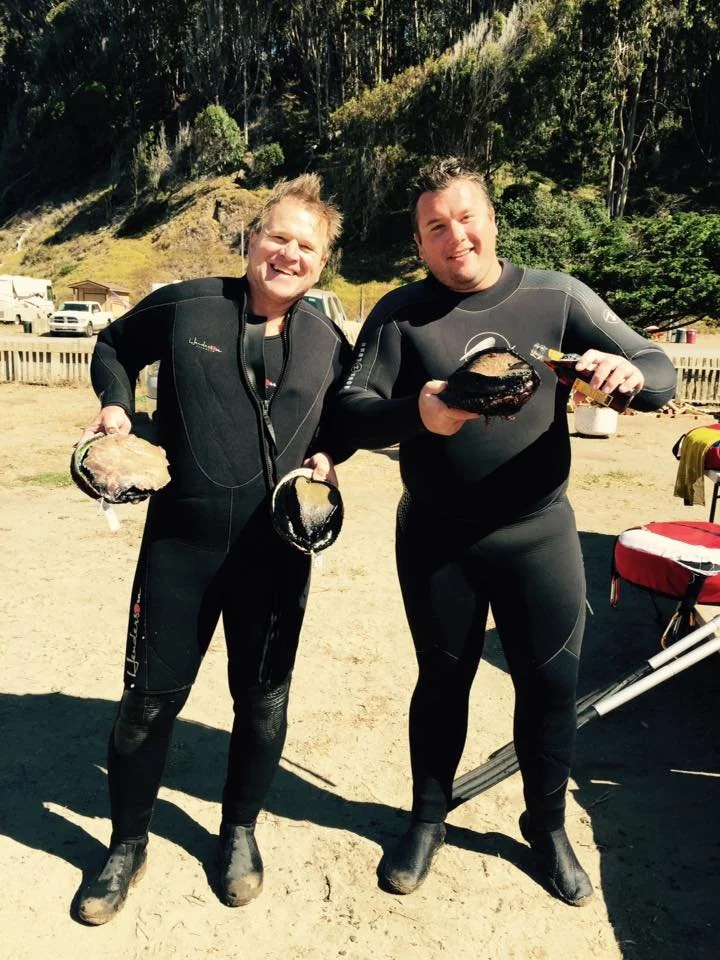 Two men in wetsuits standing outdoors, holding large pieces of cooked food, smiling at the camera. The background shows a wooden fence, parked cars, and a hillside with trees.