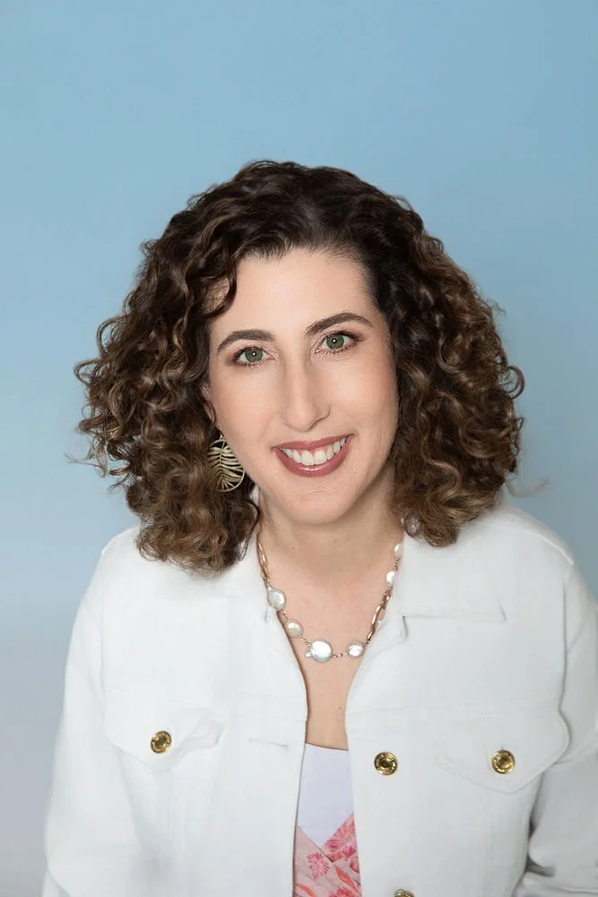 A woman with curly brown hair, green eyes, and a bright smile, wearing a white jacket with gold buttons, a pearl necklace, and gold earrings, against a light blue background.