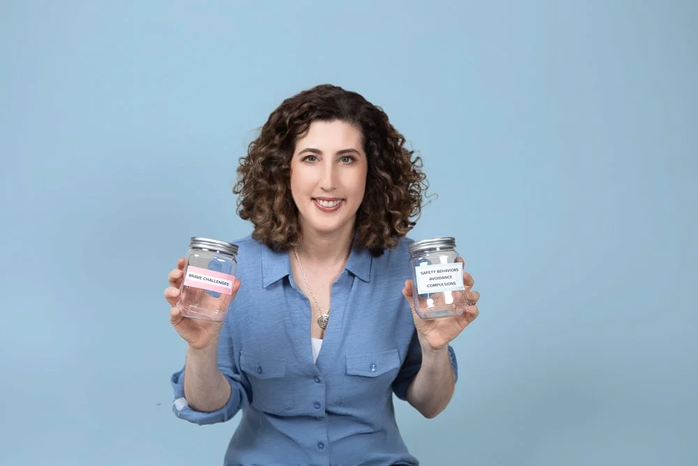 Woman holding two glass jars labeled 'Brave Challenges' and 'Safety Behaviors, Advance Compliance' against a blue background.