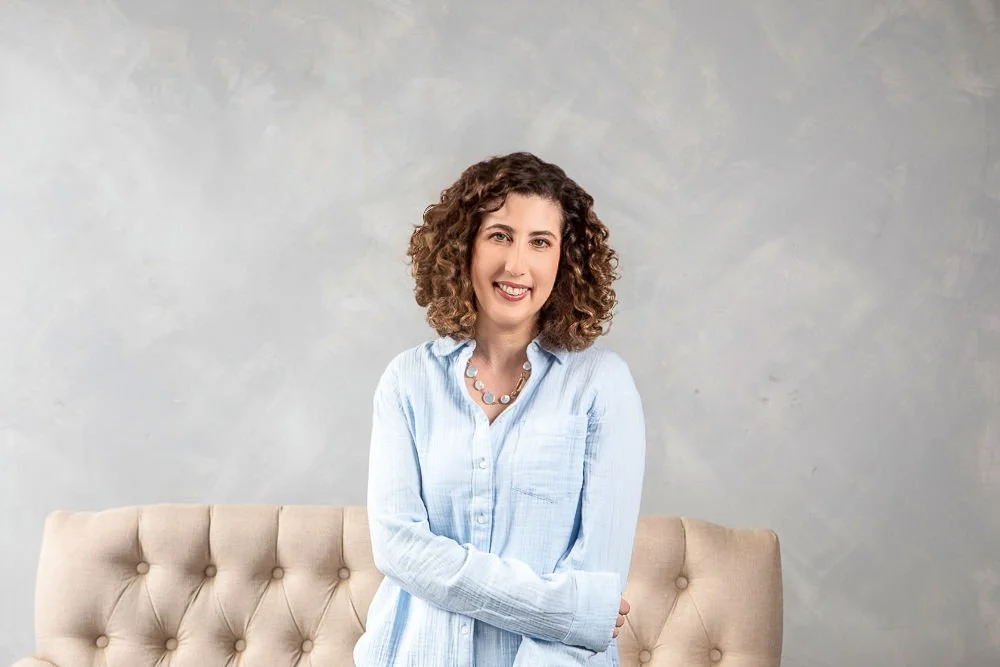 A woman with curly brown hair standing in front of a beige tufted sofa and a gray wall, smiling at the camera.