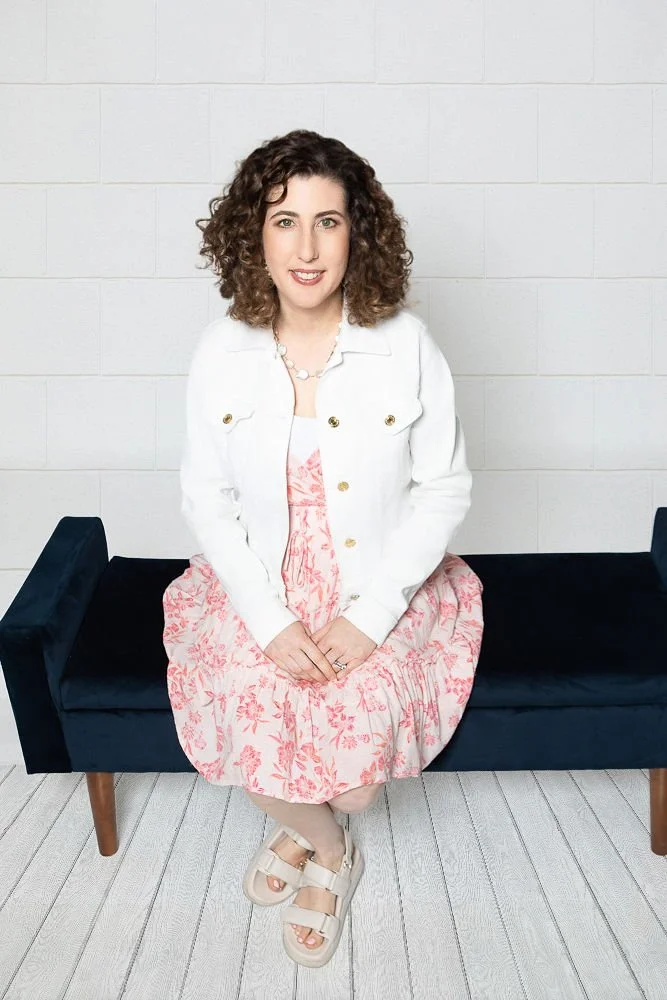A woman with curly brown hair, wearing a white jacket over a pink floral dress, sitting on a black bench against a white brick wall.