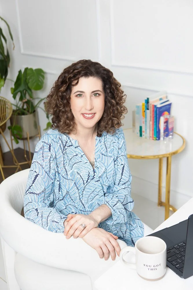 Woman with curly brown hair smiling while sitting at a white desk in a bright room, with a laptop, a mug, and a bookshelf in the background.