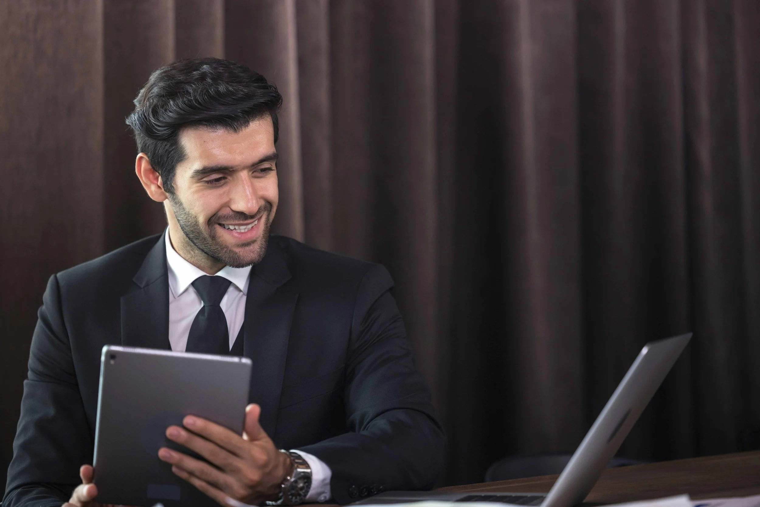 A smiling man in a business suit holding a tablet while sitting at a desk with a laptop.