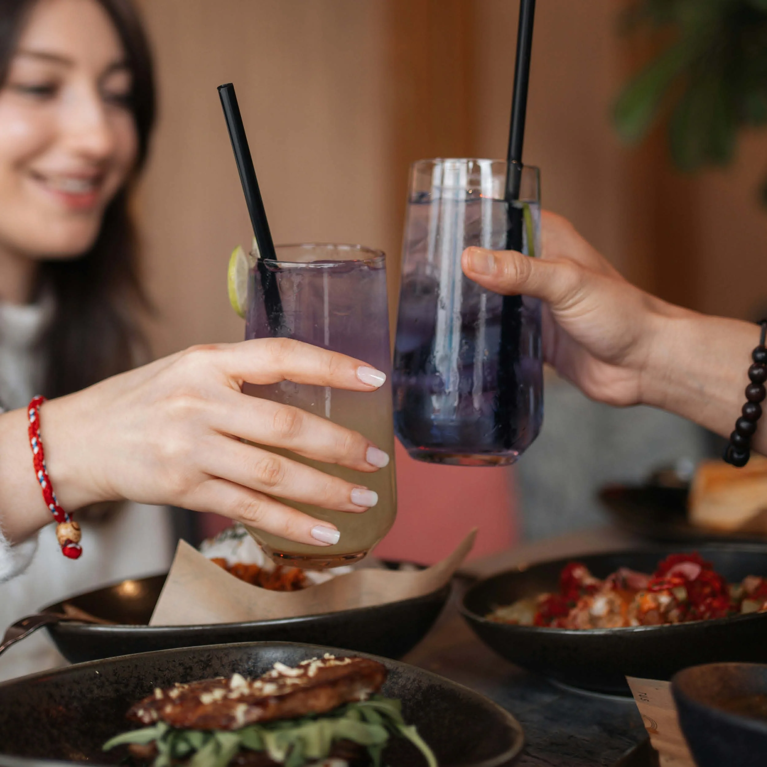 Two women are raising glasses of purple beverages with black straws in a toast, surrounded by plates of food on a table.