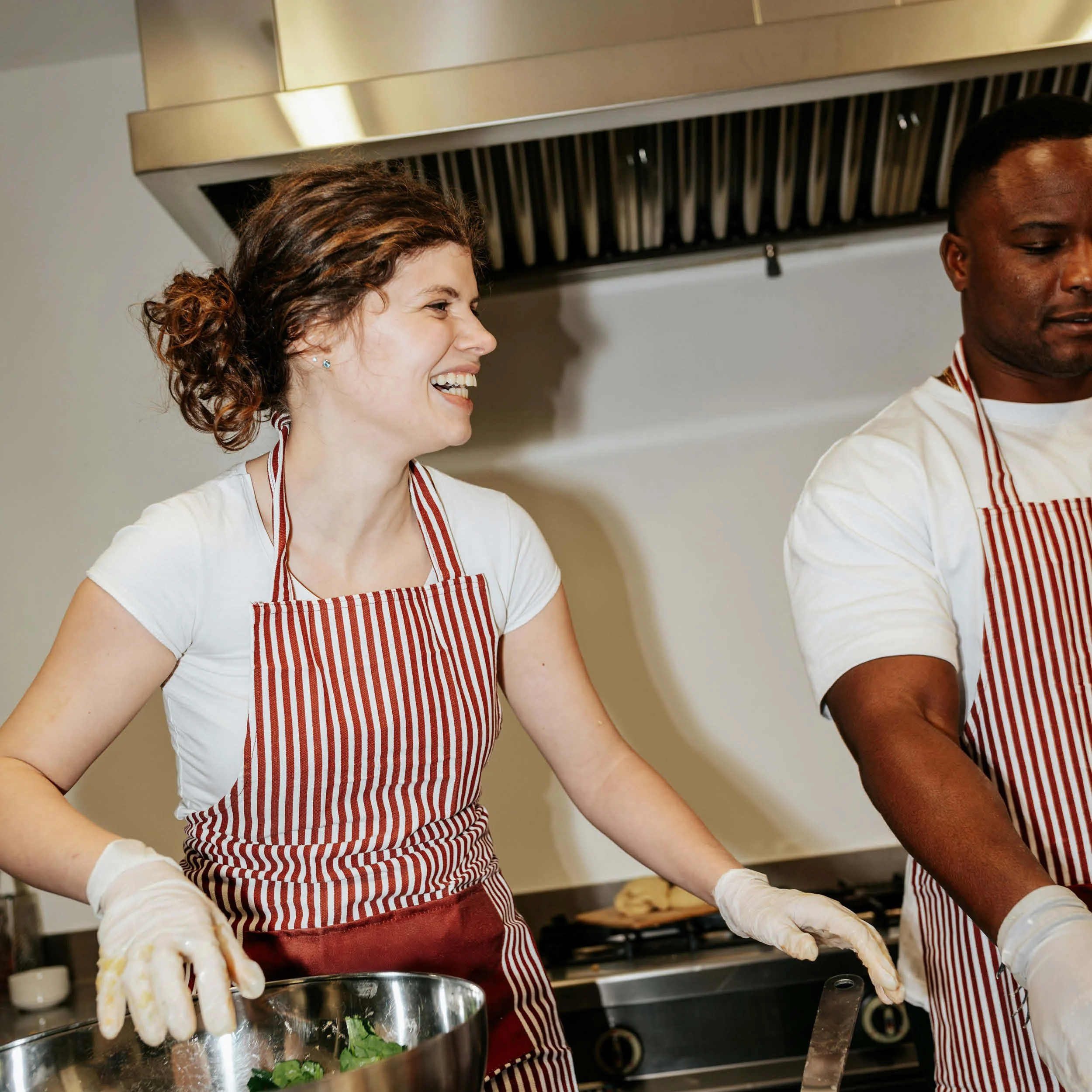Two people wearing red and white striped aprons and white gloves cooking in a kitchen, one woman laughing and the other person focused.