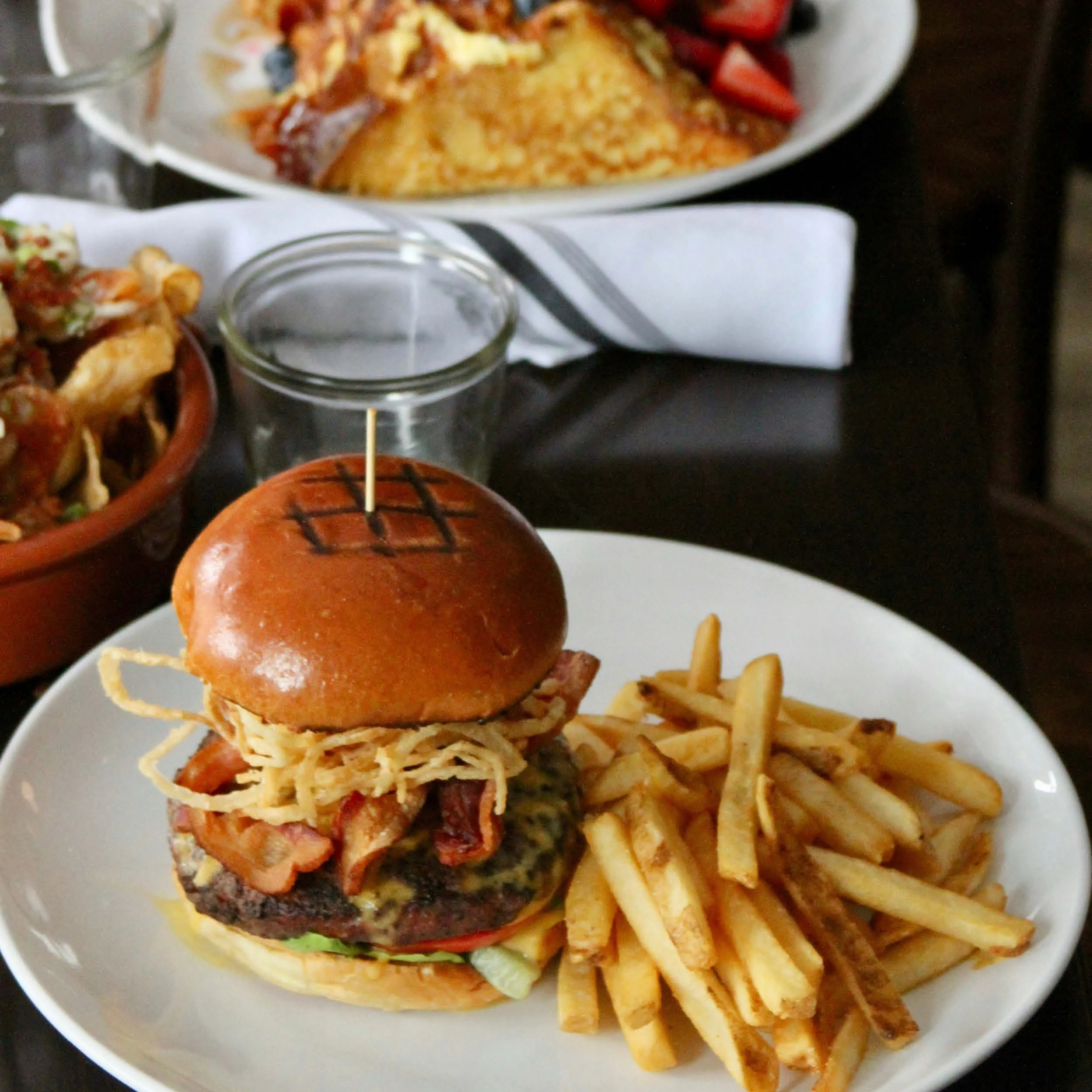 A plate of food with a cheeseburger topped with crispy fried onion strings and bacon, served with a side of French fries. In the background, there are additional dishes including a bowl of nachos with cheese, tomatoes, and other toppings, and another plate with an omelet with diced tomatoes on top.