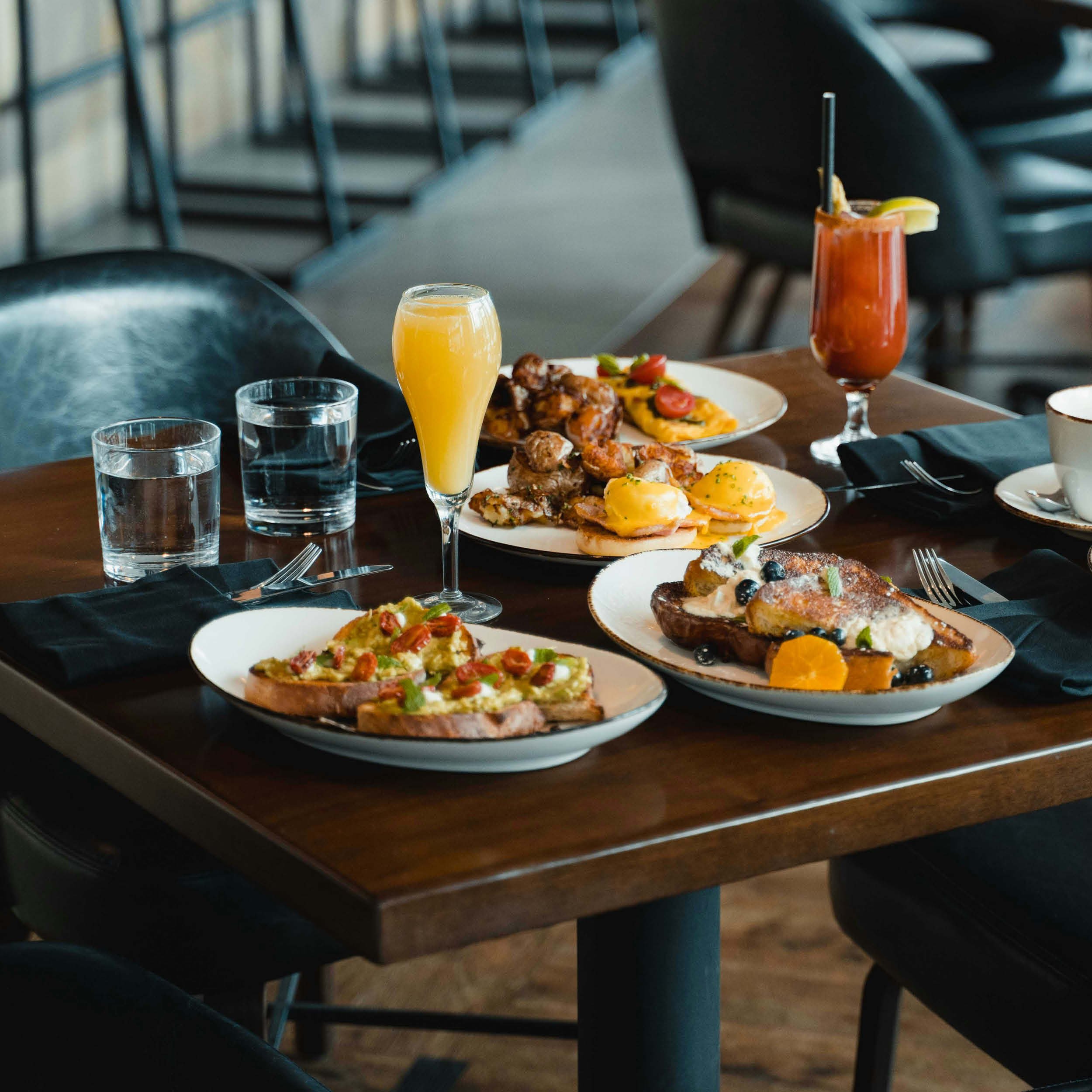 A wooden table set with various plates of breakfast food, including eggs Benedict, toast with avocado and cherry tomatoes, a plate of chicken wings, and a fruit-topped pancake. There are three glasses of water, a glass of orange juice, and a Bloody Mary cocktail with lemon and straw, in a restaurant setting with black chairs and a dark backdrop.